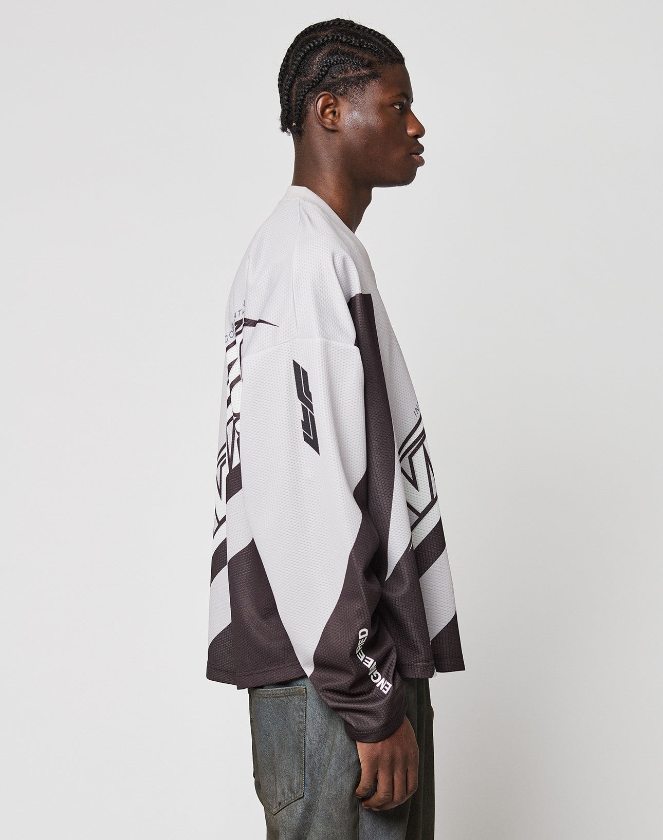 A person with braided hair stands in profile against a plain background, wearing the LIVE FAST DIE YOUNG LFDY Racetrack Jersey in black and white geometric polyester, paired with dark pants.