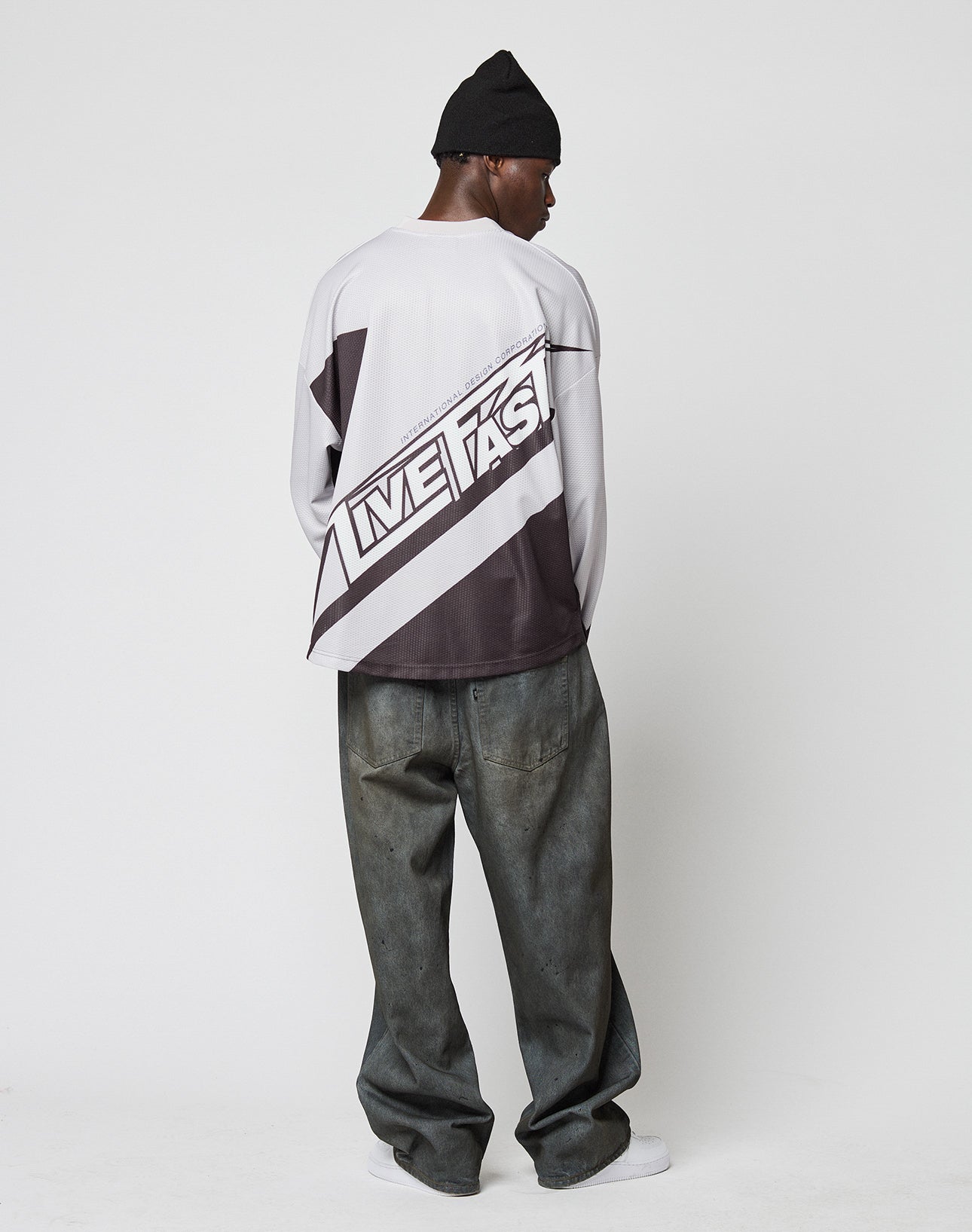 A person wearing the LFDY Racetrack Jersey by LIVE FAST DIE YOUNG, featuring an oversize fit and LIVE FAST on the back, pairs it with loose dark jeans and white sneakers while facing away from the camera against a plain white background.