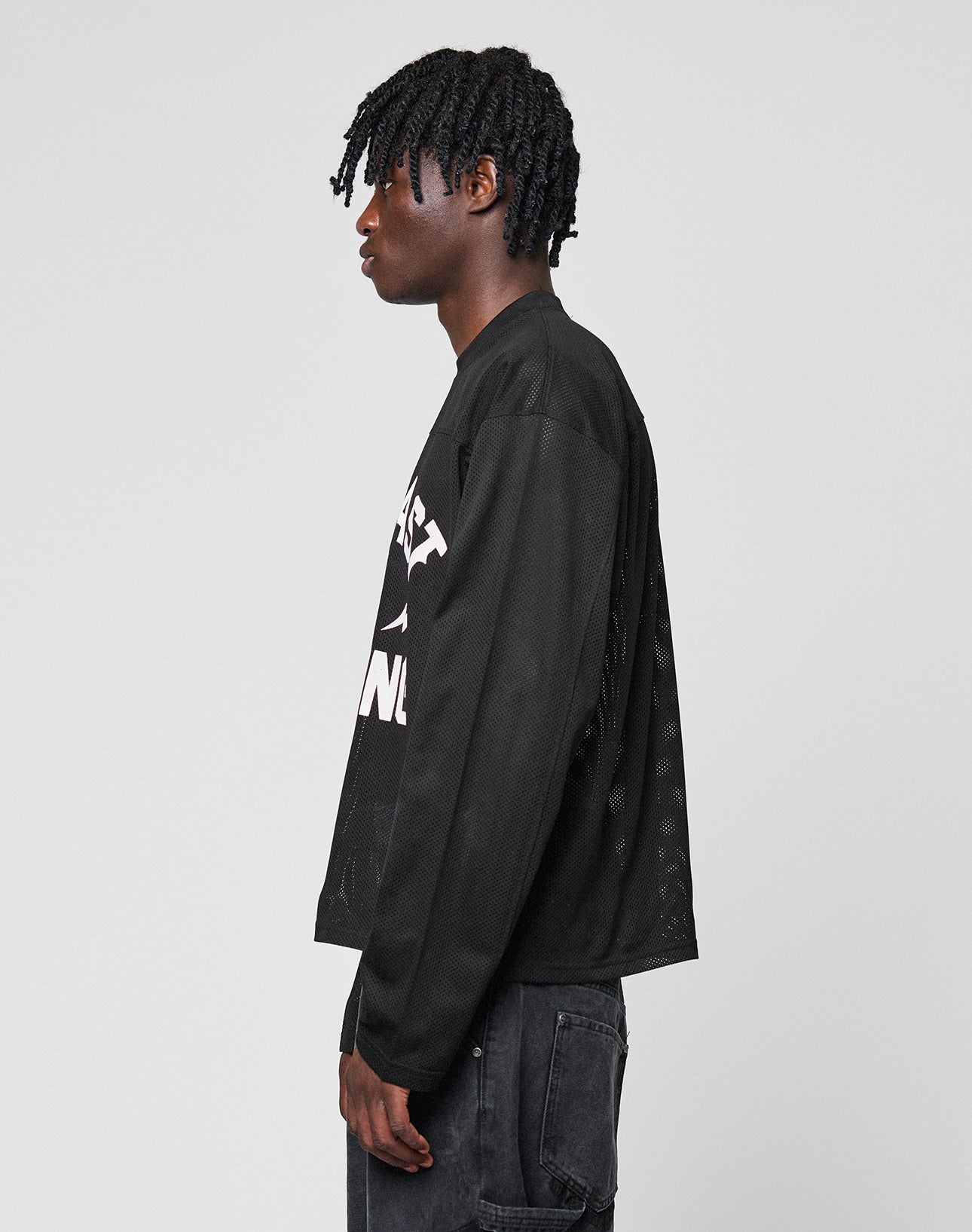 A person with short, twisted hair stands in profile wearing the LIVE FAST DIE YOUNG LFDY Flash Jersey—an oversize black long-sleeve mesh shirt with a bold front print and white text—paired with black jeans against a light gray background.