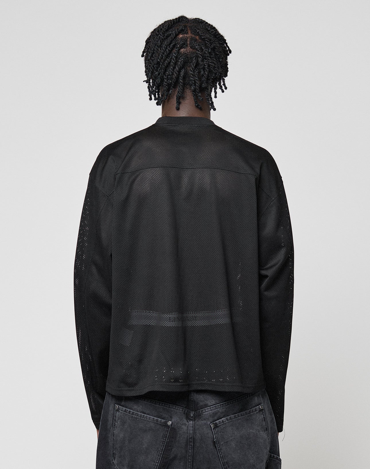 A person with short, twisted braids is seen from behind wearing the LFDY Flash Jersey by LIVE FAST DIE YOUNGāan oversize-fit, long-sleeve black mesh topāpaired with faded black jeans against a plain light gray background.