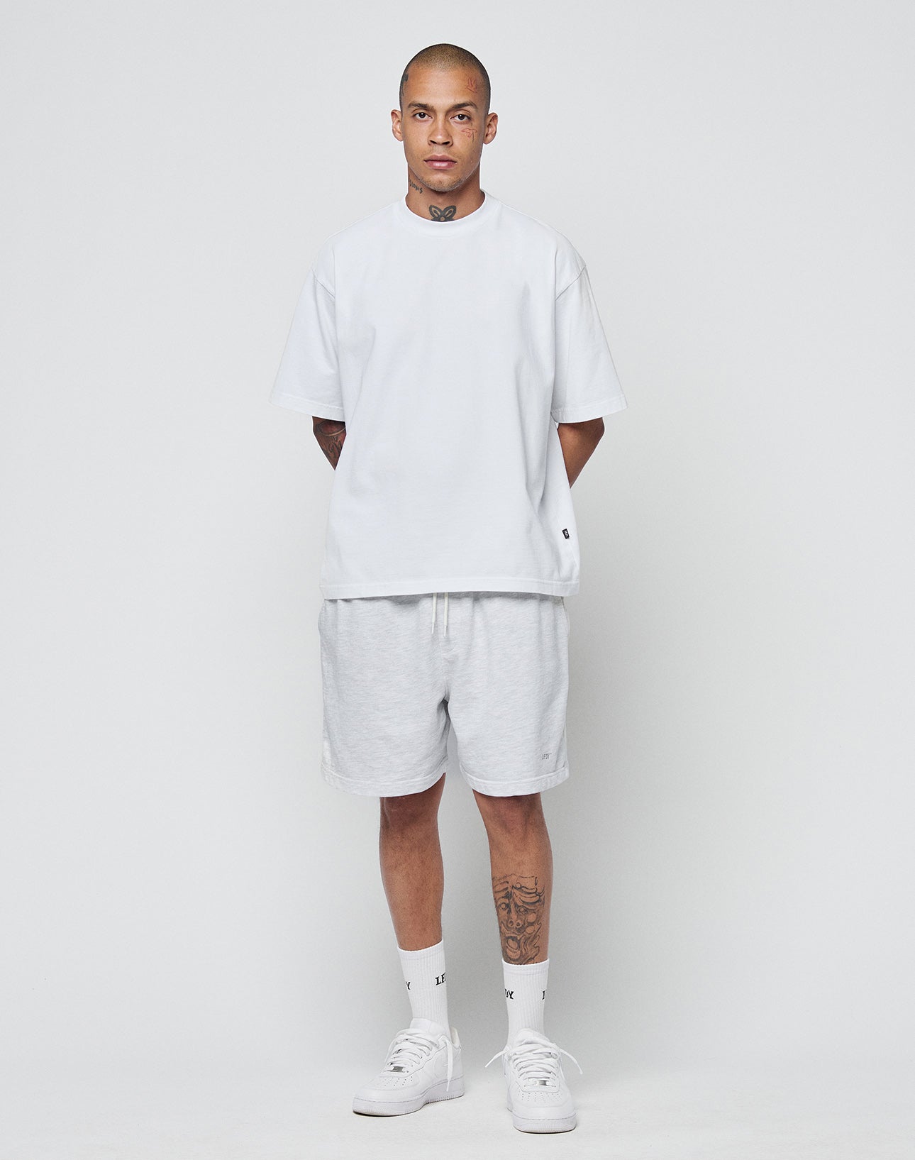 A person with short hair and visible tattoos models the LIVE FAST DIE YOUNG Basic Heavy Tee (100% Baumwolle) in an Oversize Fit, paired with light gray shorts, white socks, and sneakers against a plain white background.