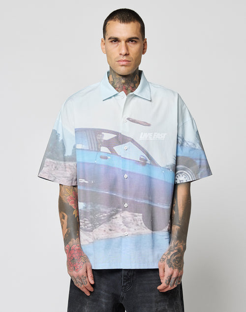 A tattooed person wears the LIVE FAST DIE YOUNG LFDY Crash Kid Shirt—an oversize, light blue short-sleeve button-up with a large blue car graphic and LIVE FAST text—standing against a plain white background.