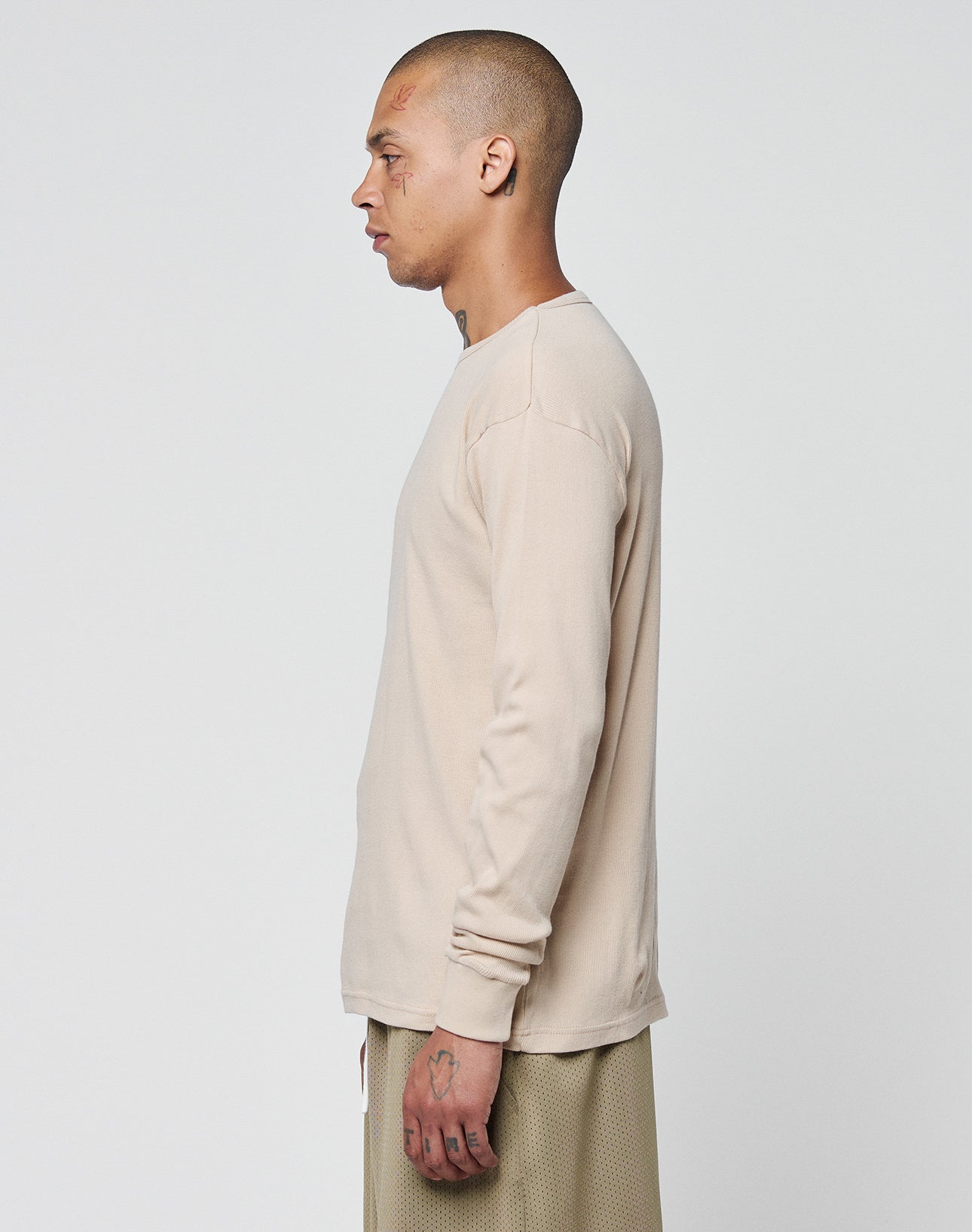A person with short hair and visible tattoos stands in profile, facing left, wearing a beige LIVE FAST DIE YOUNG Basic Regular Rib Longsleeve and light brown textured pants against a plain white background.