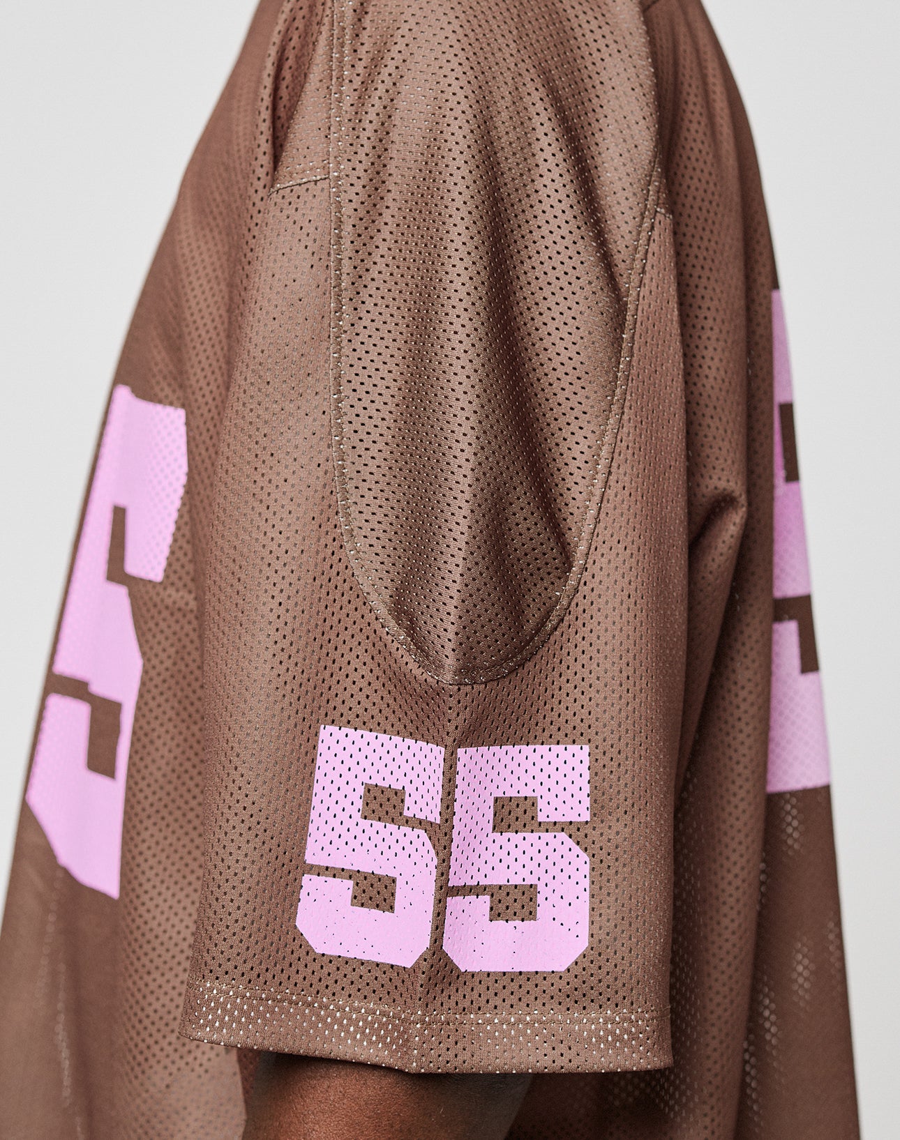 A side view of a person wearing the LIVE FAST DIE YOUNG LFDY Mesh Jersey, an oversized brown sports top featuring large pink number 55 on the sleeve and back.