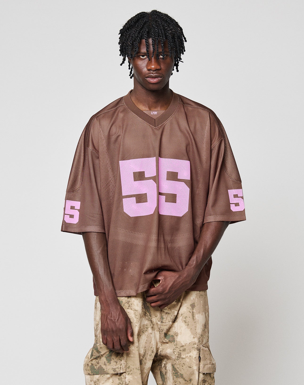 A young man with short dreadlocks wears the LFDY Mesh Jersey by LIVE FAST DIE YOUNG, featuring a loose brown mesh design with a V-neck and pink number 55, paired with beige camo cargo pants against a plain light gray background.