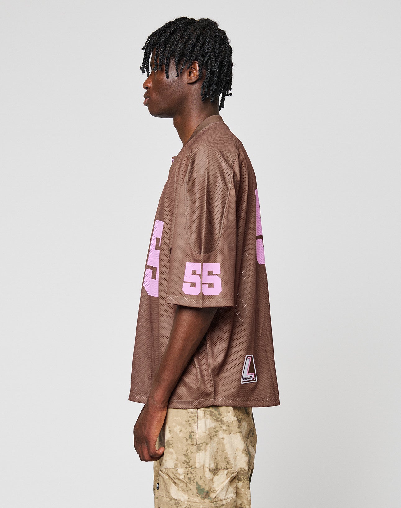 A person with short, twisted hair wears the LIVE FAST DIE YOUNG LFDY Mesh Jersey in brown—oversize fit with a pink 55 on the sleeve, V-Ausschnitt—plus light camouflage pants, standing in profile against a plain background.