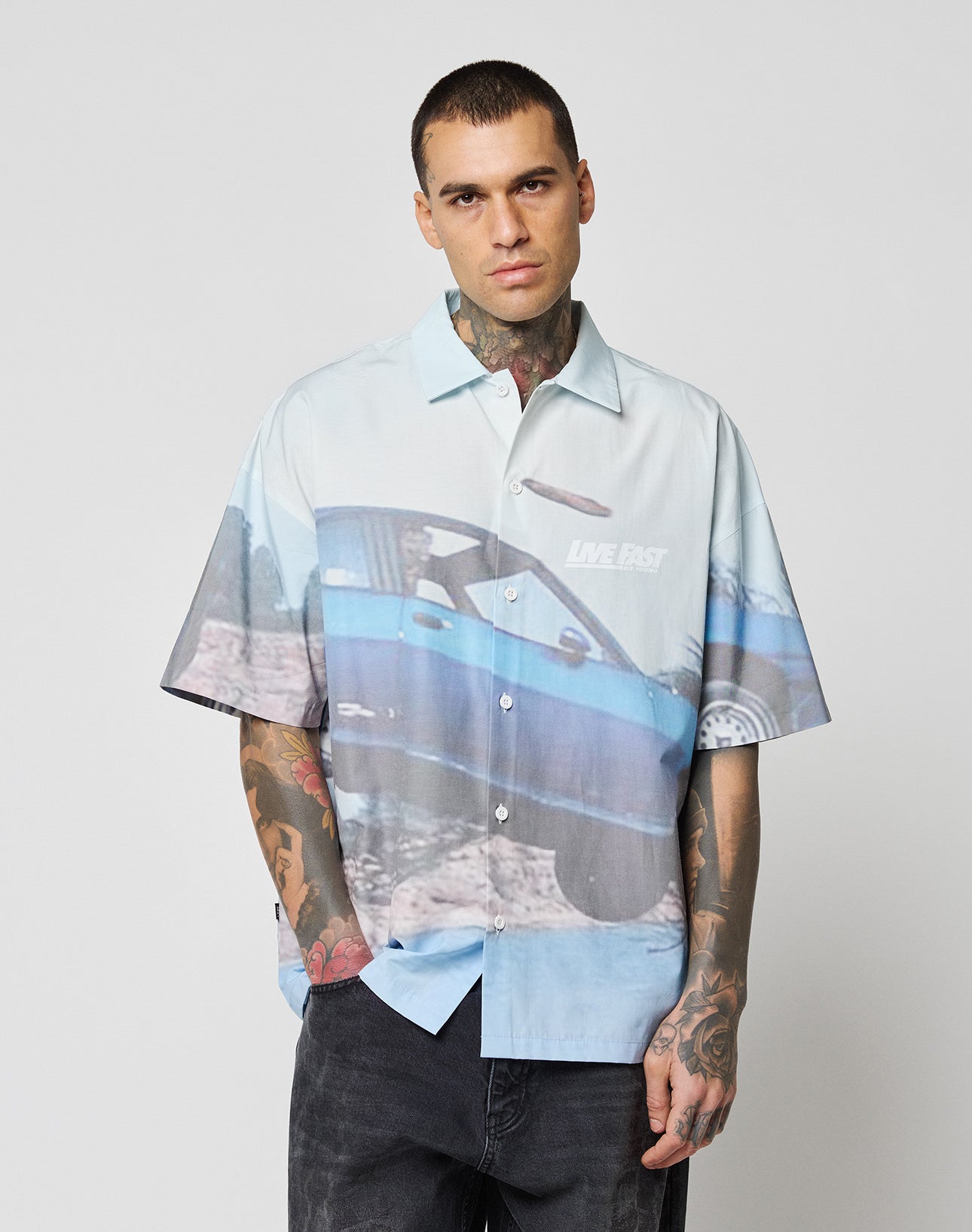 A man with tattoos and short dark hair wears the LIVE FAST DIE YOUNG LFDY Crash Kid Shirt, an oversized light blue top featuring a large blue car graphic and LIVE FAST text, standing against a plain light background.