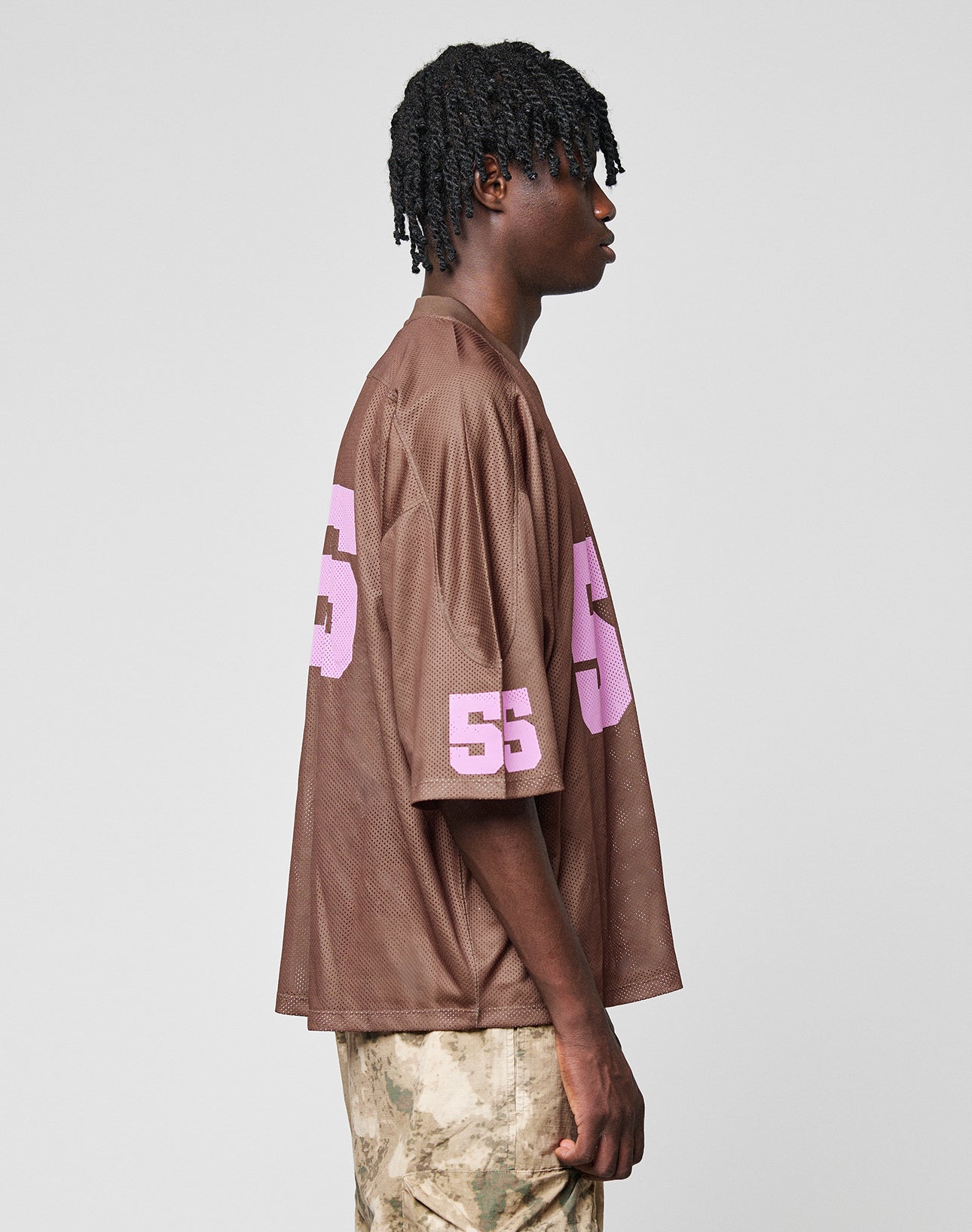 A person stands in profile wearing the LIVE FAST DIE YOUNG LFDY Mesh Jersey—a brown sports top with a V-neck and pink number 55 on the sleeve and back—paired with beige camo pants against a plain light background.