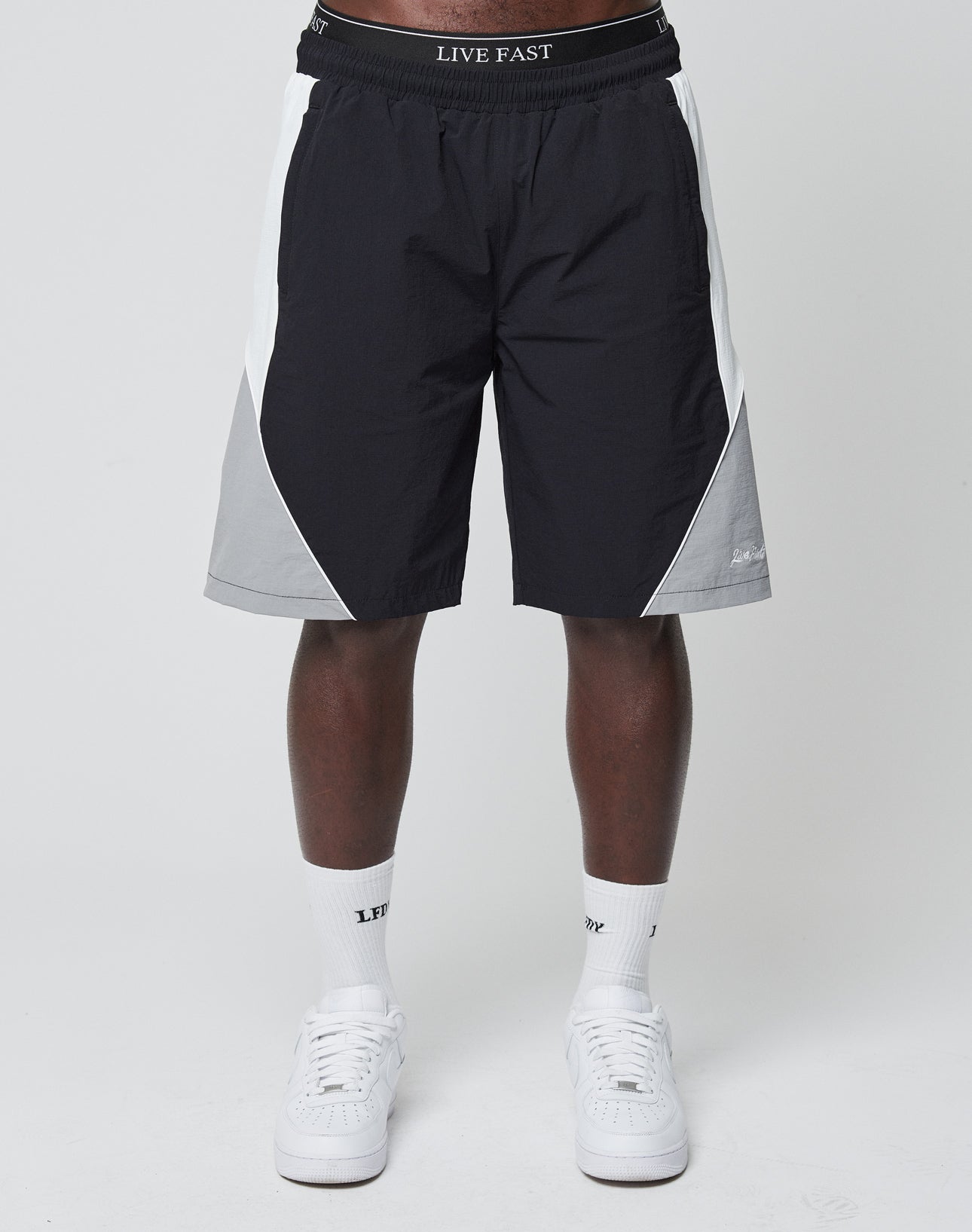 A person wears LIVE FAST DIE YOUNG Basic Nylon Trackshorts in black, white, and gray with an elastic waistband, paired with white crew socks and sneakers, standing against a plain light background.