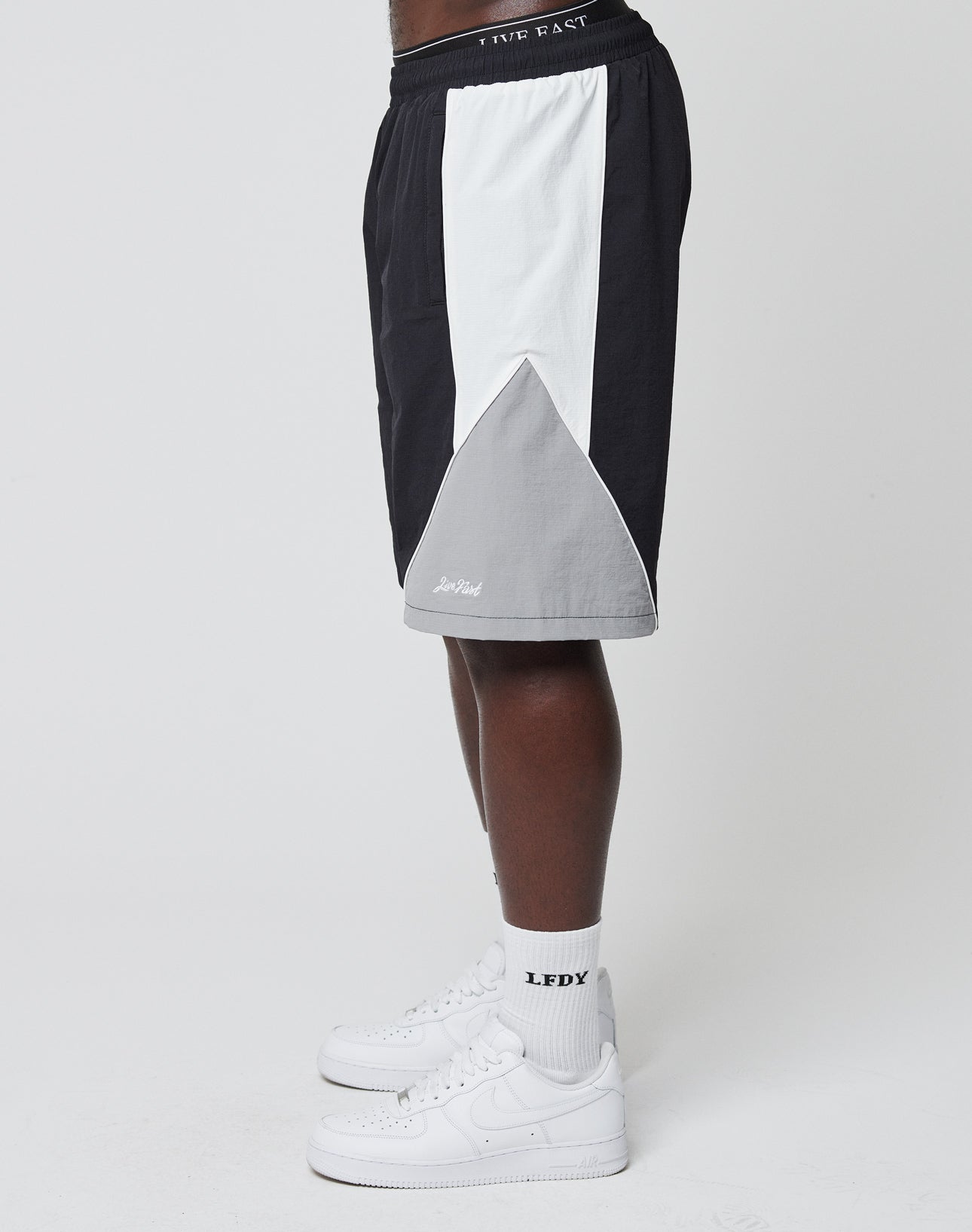 A person stands sideways against a plain white background wearing LIVE FAST DIE YOUNG Basic Nylon Trackshorts in black, white, and gray with an elastic waistband, paired with white LFDY socks and white Nike Air Force 1 sneakers.