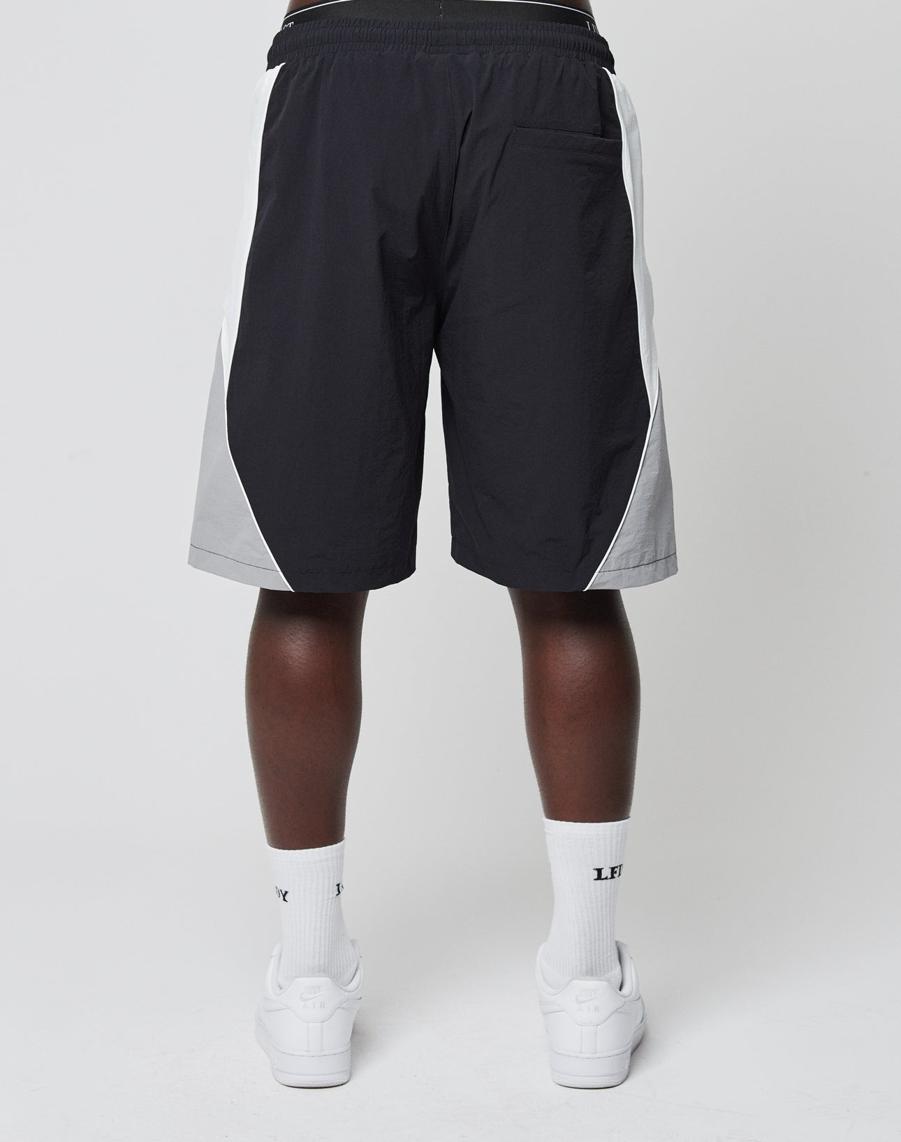 A person faces away from the camera wearing LIVE FAST DIE YOUNG Basic Nylon Trackshorts, white crew socks with LFE logo-stick detail, and white sneakers against a plain light background.