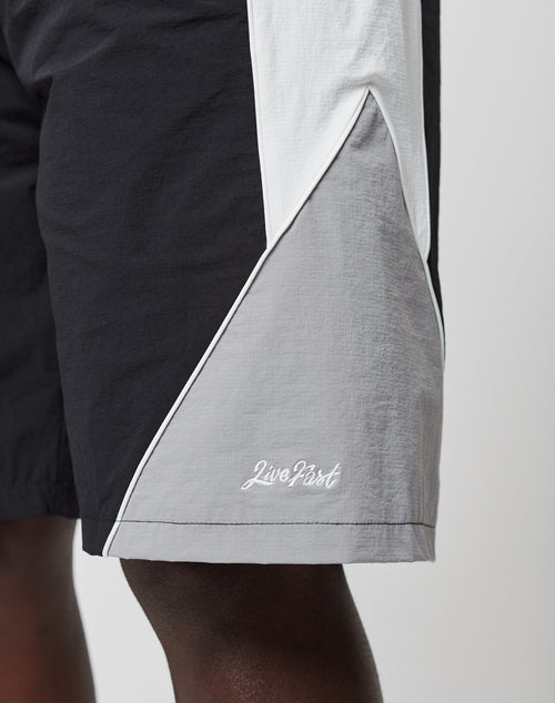 Close-up of a person wearing LIVE FAST DIE YOUNG Basic Nylon Trackshorts in black, white, and gray with Live Fast embroidery on the lower leg. Features an elastic waistband. Only part of the leg and shorts are visible.