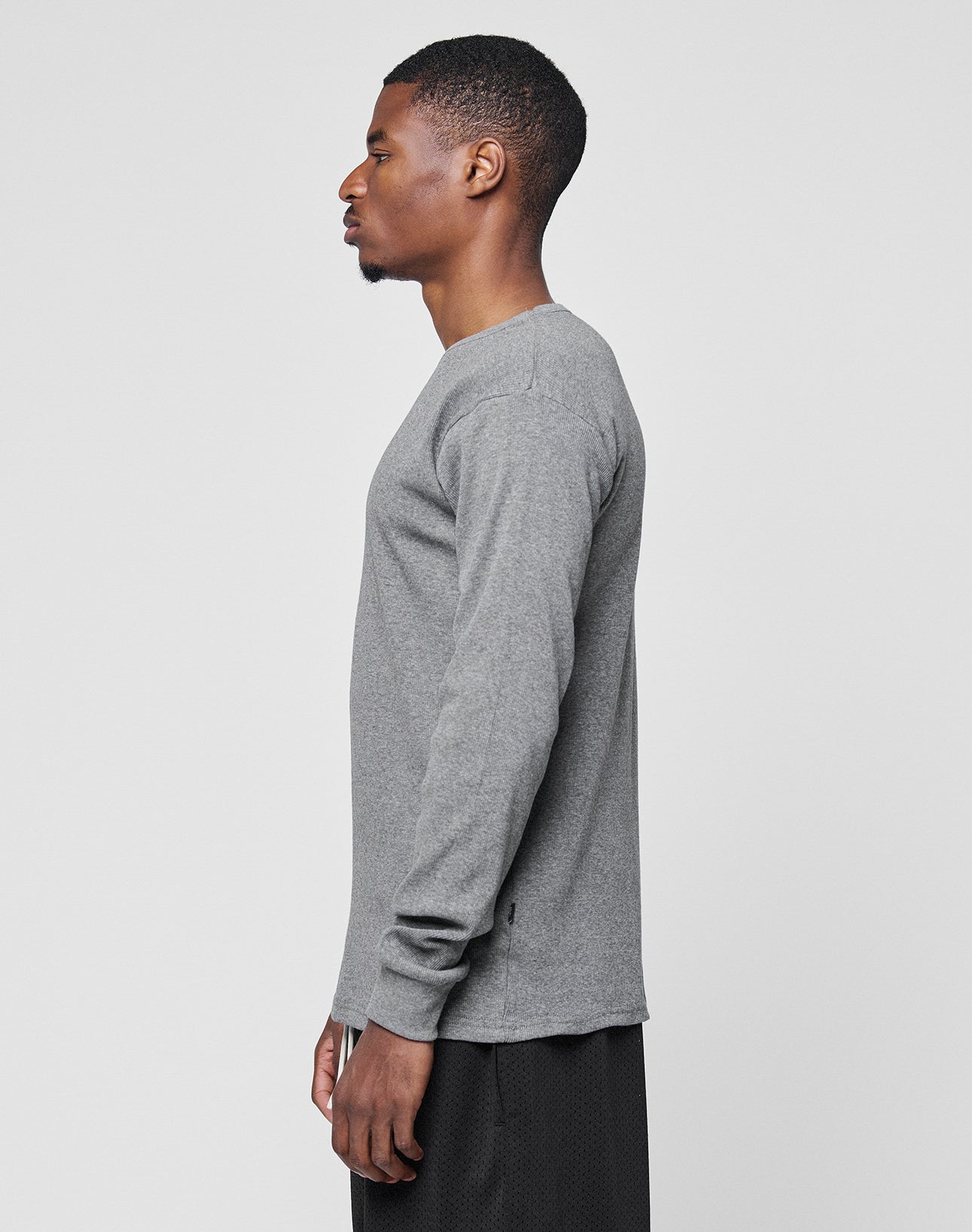 A man stands in profile facing left, wearing the LIVE FAST DIE YOUNG Basic Regular Rib Longsleeve in gray and black shorts against a plain light gray background.