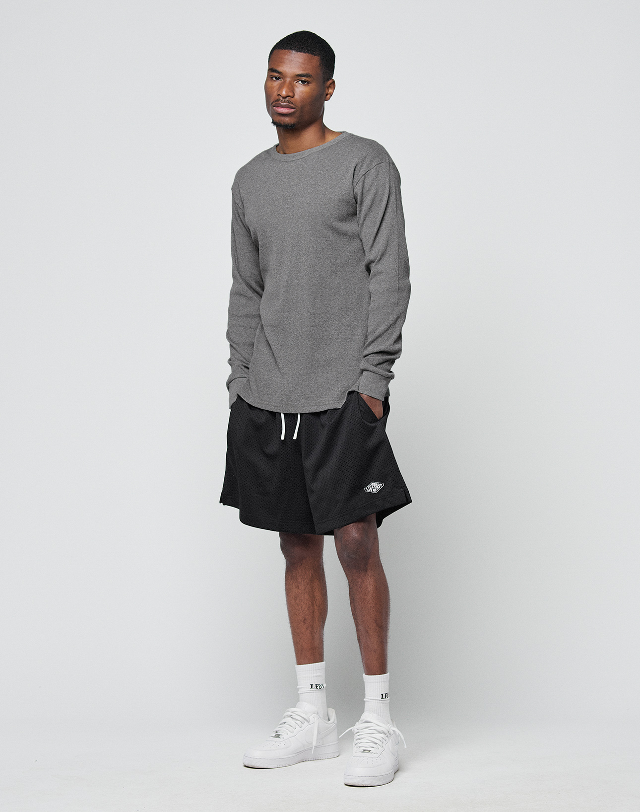 A man stands against a white background, wearing a LIVE FAST DIE YOUNG Basic Regular Rib Longsleeve in gray, black drawstring shorts, white crew socks, and white sneakers. His hands are in his pockets and he has a neutral expression.