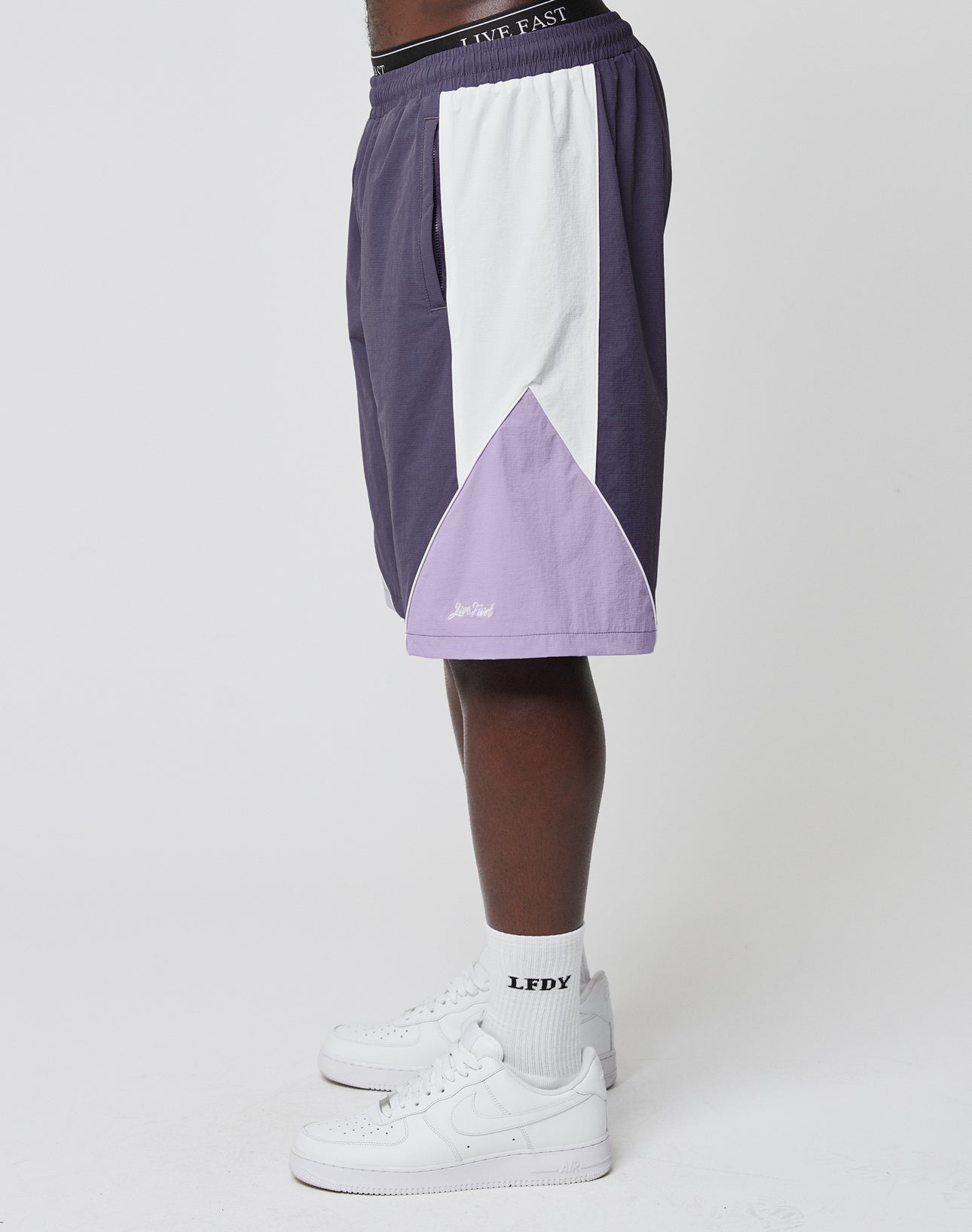 A person stands sideways wearing LIVE FAST DIE YOUNG Basic Nylon Trackshorts in purple, white, and lavender color-block with an elastic waistband, paired with LEDX white socks and white sneakers against a plain white background.