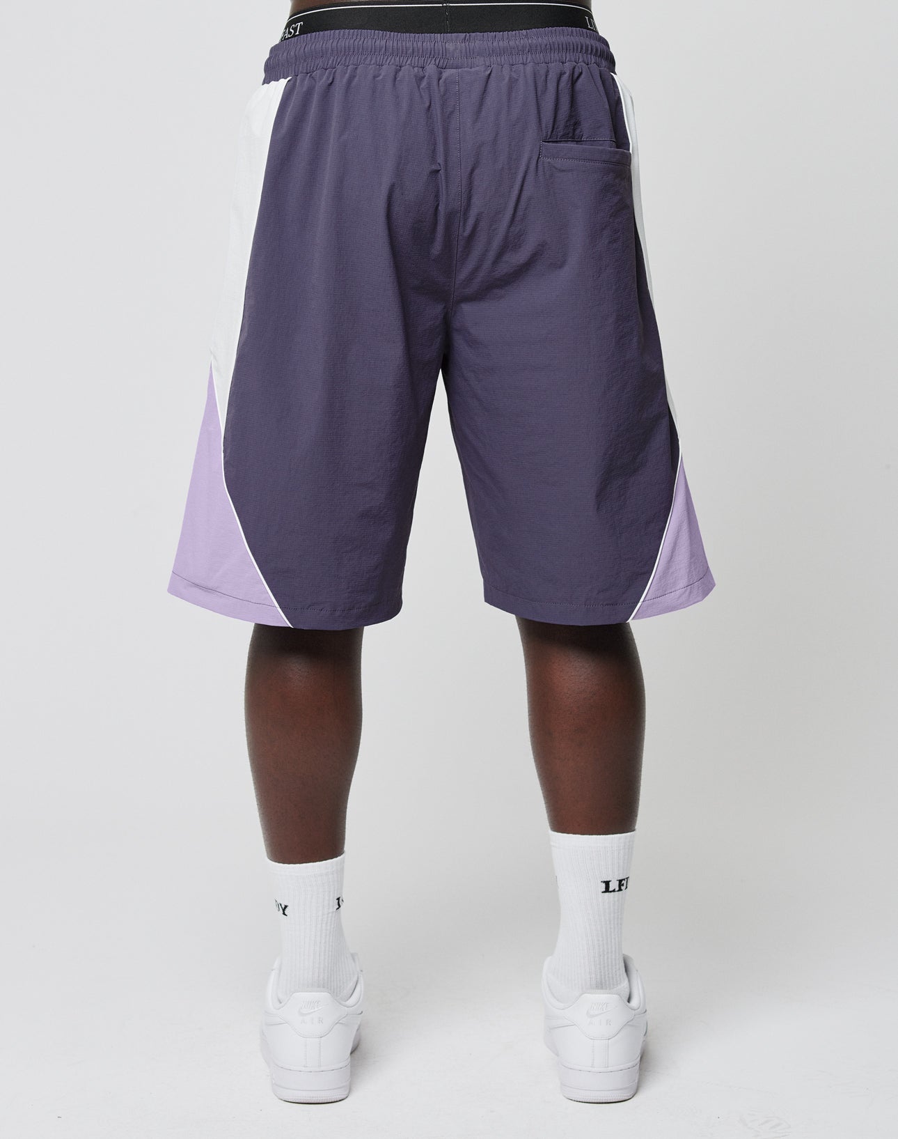 A person faces away, wearing LIVE FAST DIE YOUNG Basic Nylon Trackshorts in purple and white with an elastic waistband, paired with white LFC socks and white sneakers, standing against a plain white background.