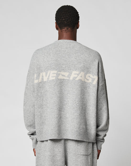 A person faces away in a light gray, oversize Live Fast Boucle Sweater by LIVE FAST DIE YOUNG, featuring “LIVE FAST” in white on the back. Made from 100% polyester and paired with matching gray pants.