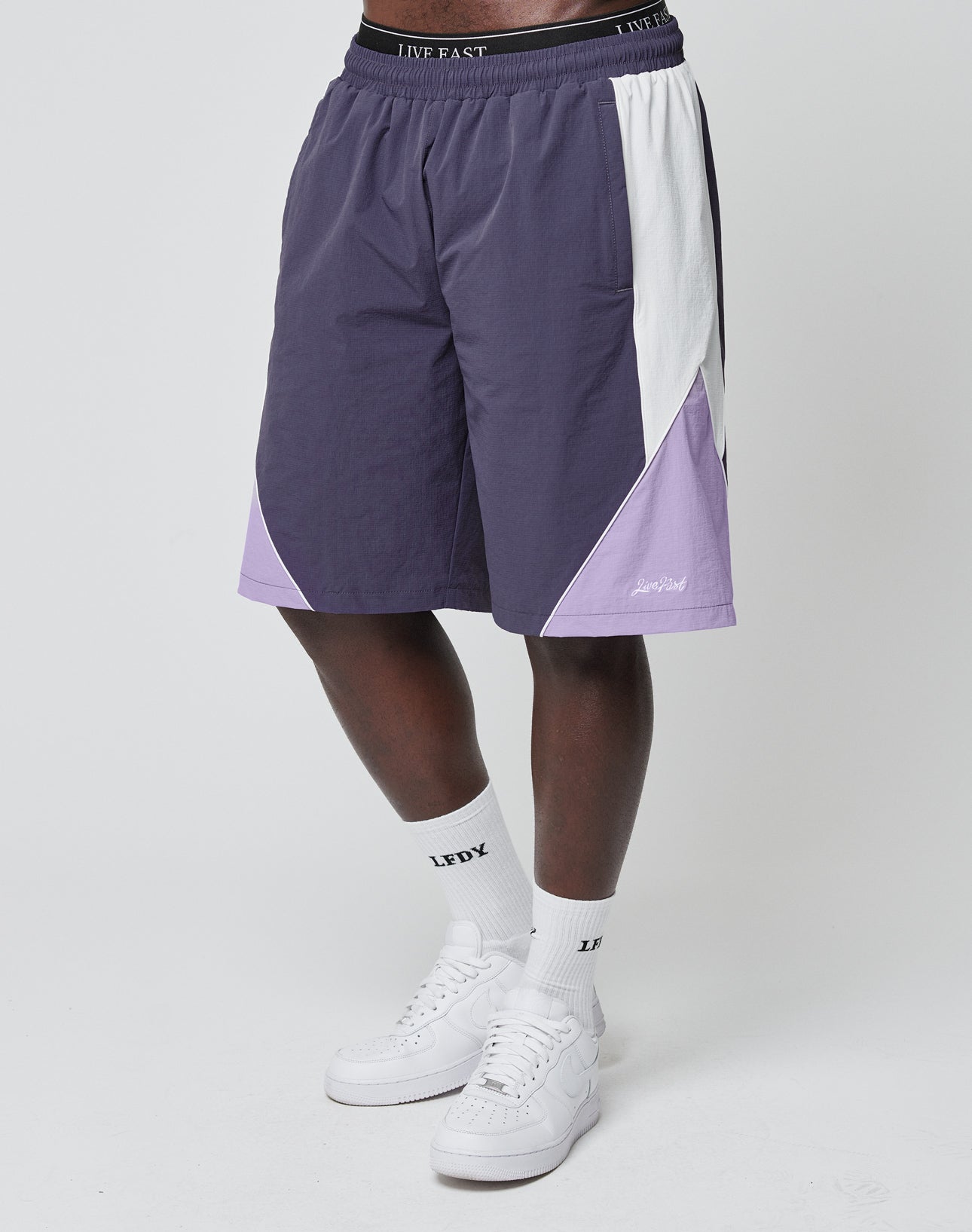 A person wears LIVE FAST DIE YOUNG Basic Nylon Trackshorts in purple and white with an elastic waistband, paired with LFYD logo white socks and white sneakers, standing against a plain white background.
