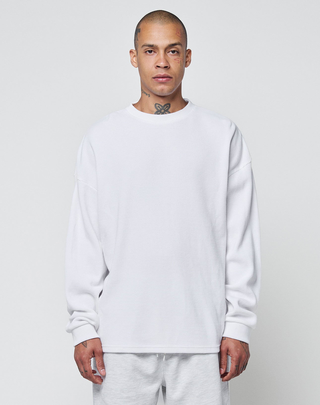 A person with a shaved head and visible neck tattoo faces forward against a plain light gray background, wearing the LIVE FAST DIE YOUNG Blank Waffle Longsleeve in white with Ripp-Bündchen and matching white pants.