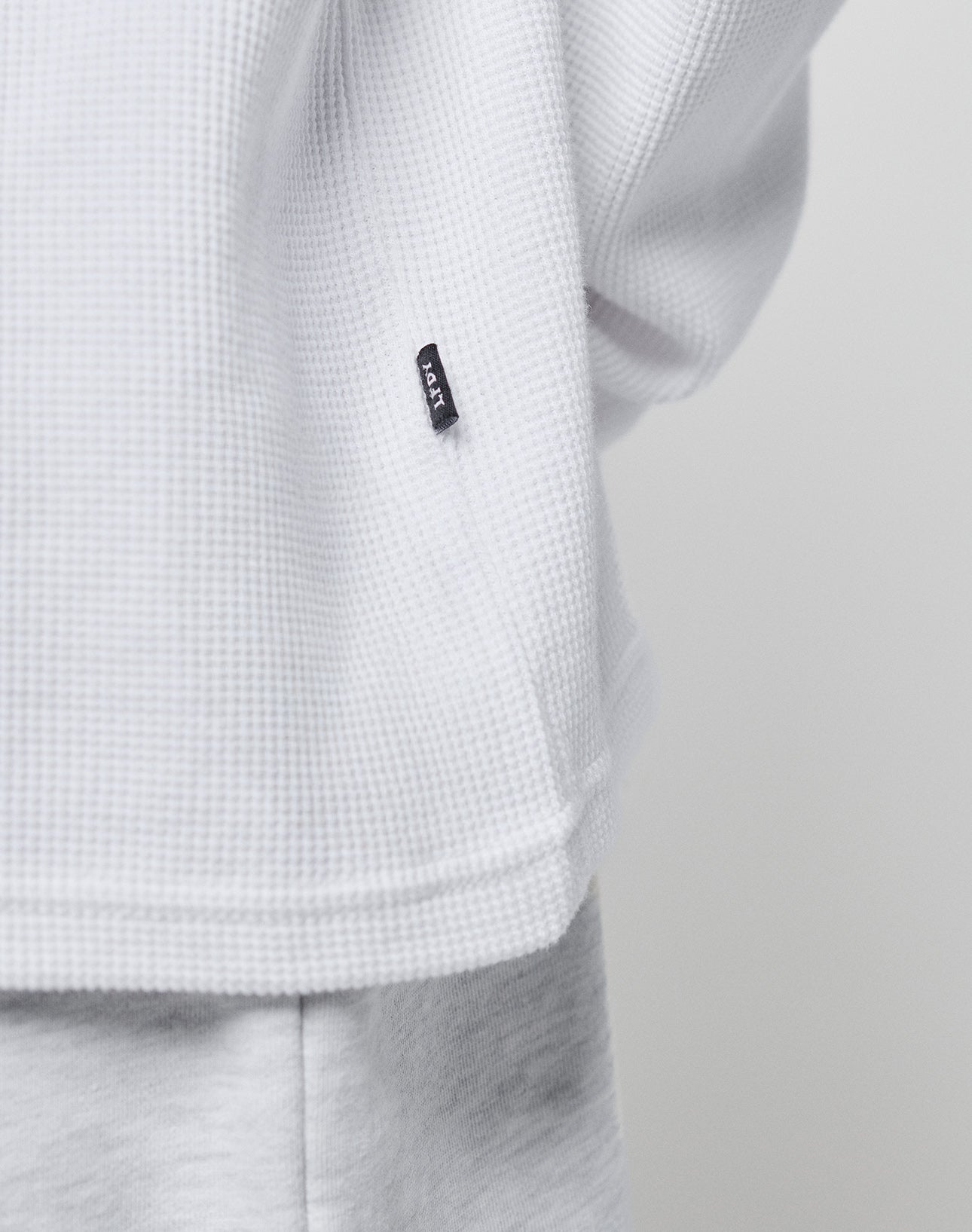 Close-up of the LIVE FAST DIE YOUNG Blank Waffle Longsleeve in white, showing its textured waffle-knit fabric, Ripp-Bündchen at the hem, and a glimpse of light gray pants.