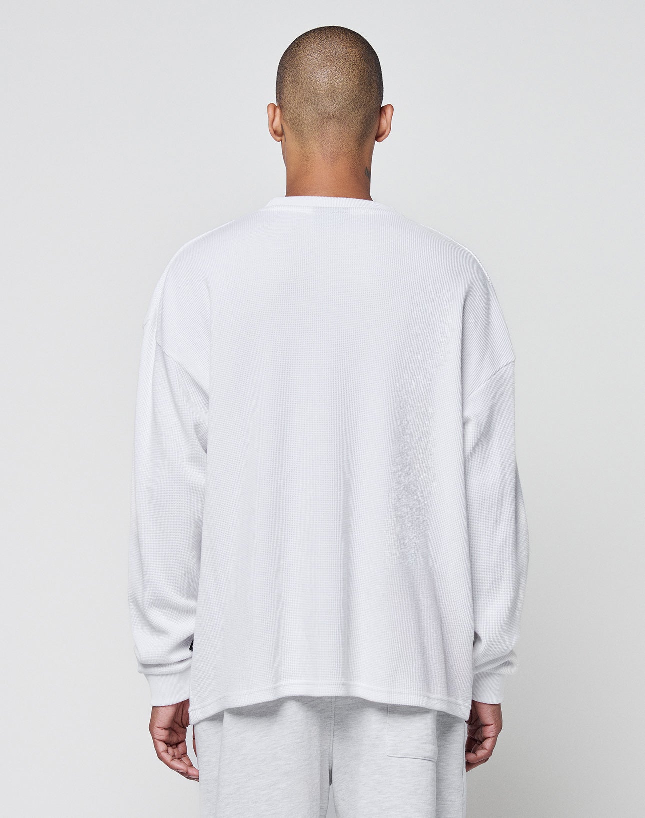 A person with a shaved head faces away from the camera, wearing the LIVE FAST DIE YOUNG Blank Waffle Longsleeve in an oversize fit and light-colored pants, set against a plain light gray background.
