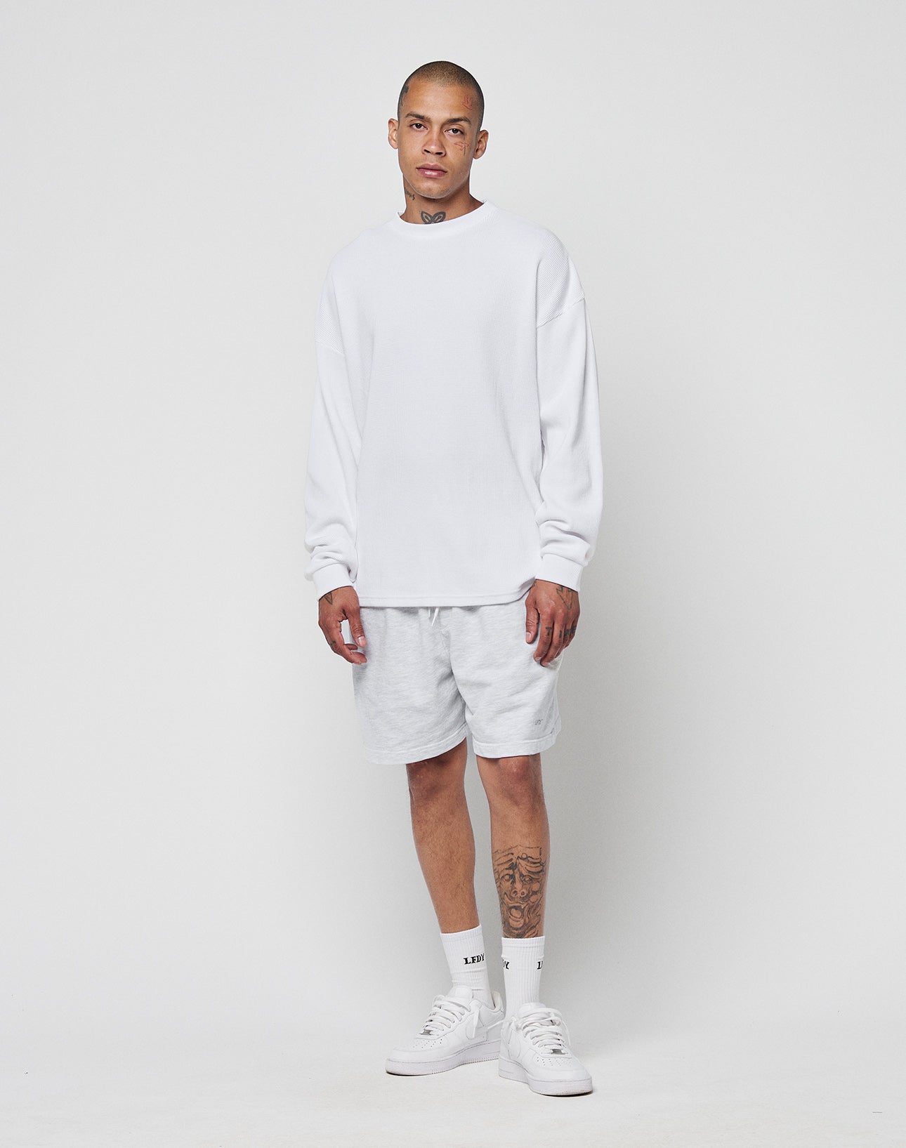 A person with neck and left leg tattoos stands against a plain white background, wearing a LIVE FAST DIE YOUNG Blank Waffle Longsleeve (Oversize Fit), light gray shorts, white socks, and white sneakers.
