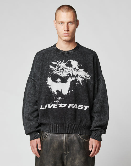 A person with short hair wears the LFDY Savior Knit Sweater by LIVE FAST DIE YOUNG—an oversized dark sweater with a bold abstract white graphic and LIVE FAST text, standing against a plain light background.