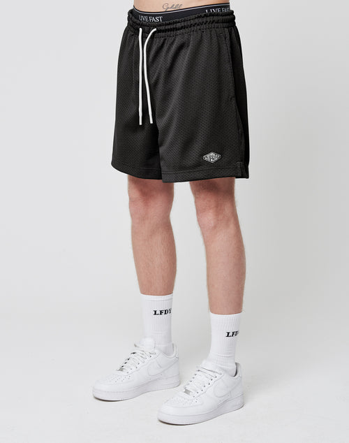 Wearing LIVE FAST DIE YOUNG Basic Mesh Shorts in black with a white drawstring and logo patch, paired with white LFDY crew socks and Nike sneakers, standing against a plain white background. Upper body is not visible.