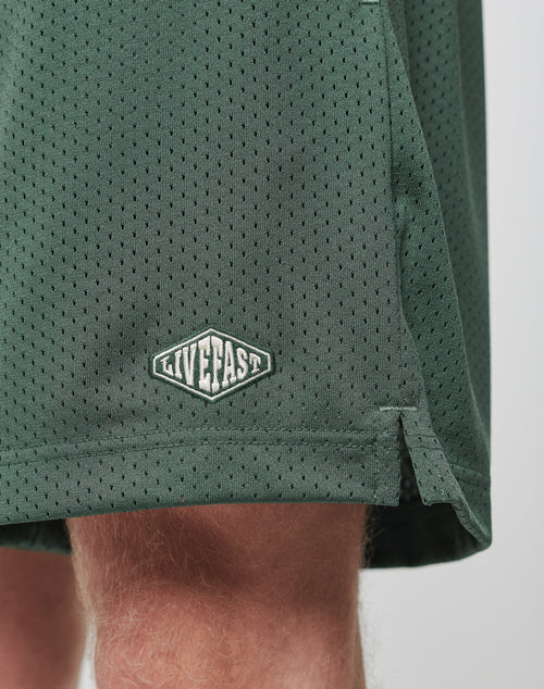 Close-up of the lower part of green Basic Mesh Shorts by LIVE FAST DIE YOUNG, featuring an elastic waistband and a small embroidered LIVE FAST logo patch. Made from polyester mesh and shown worn by someone with light skin.