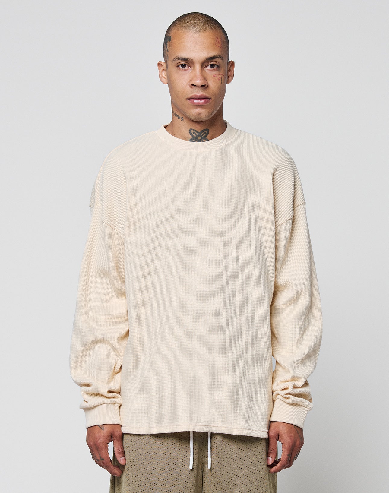 A person with a shaved head and visible neck tattoo wears the LIVE FAST DIE YOUNG Blank Waffle Longsleeve, an oversized cream 100% Baumwolle shirt, with tan shorts, standing against a plain light gray background.