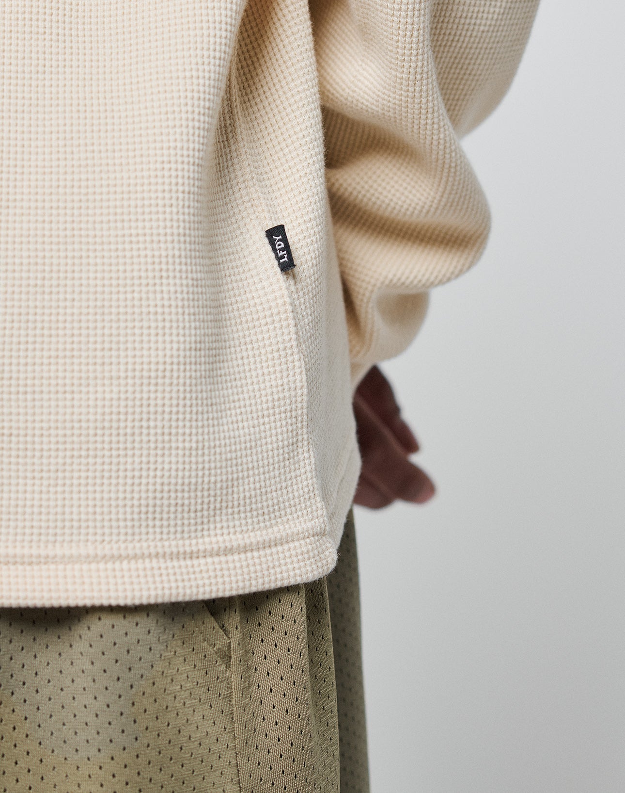 A close-up of a person wearing the LIVE FAST DIE YOUNG Blank Waffle Longsleeve in cream 100% Baumwolle, oversize fit with a small black ADAY label, paired with olive green perforated pants; one hand is partially visible in the background.