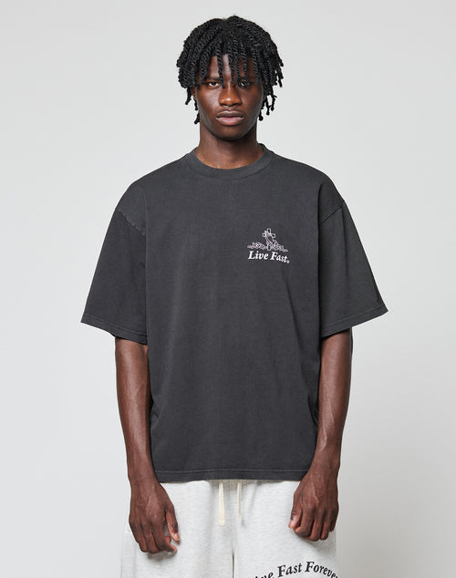 A person with short, twisted braids wears the LIVE FAST DIE YOUNG LFDY Praying Tee, a loose dark shirt featuring Live Fast text, paired with light shorts displaying Live Fast Forever on the waistband, against a plain background.