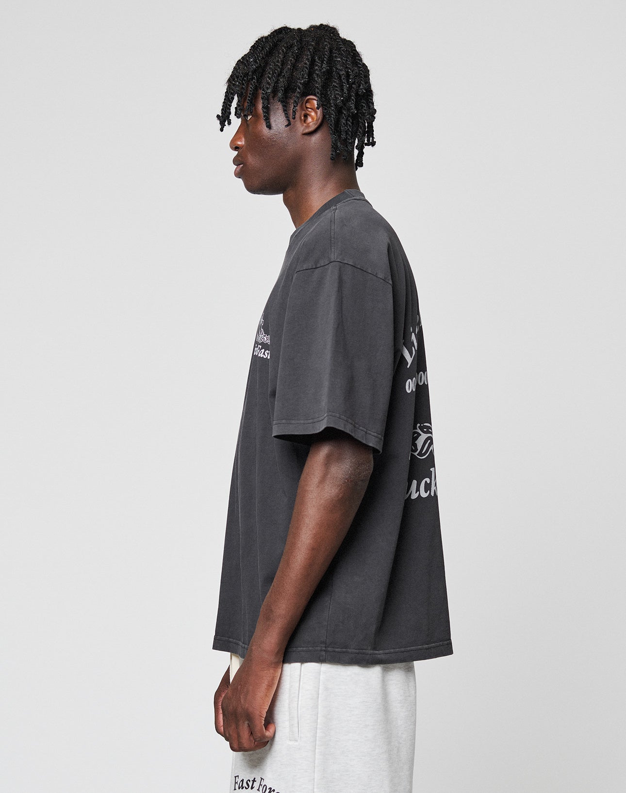 A young man with short twists stands in profile, wearing the LIVE FAST DIE YOUNG LFDY Praying Tee—an oversize black 100% Baumwolle T-shirt with bold front and back print—paired with light gray shorts on a plain light gray background.