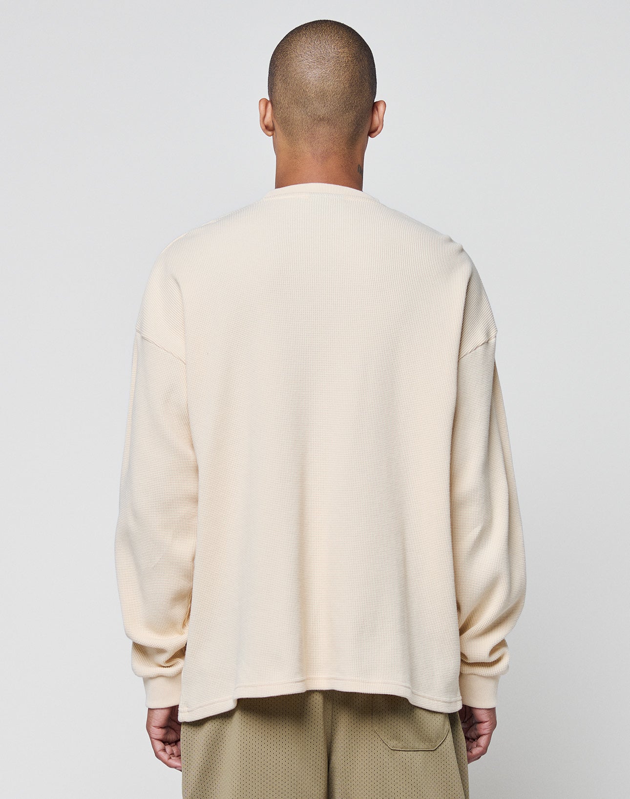 A person with a shaved head is shown from behind, wearing the LIVE FAST DIE YOUNG Blank Waffle Longsleeve in off-white 100% Baumwolle and light brown mesh shorts against a plain light background.