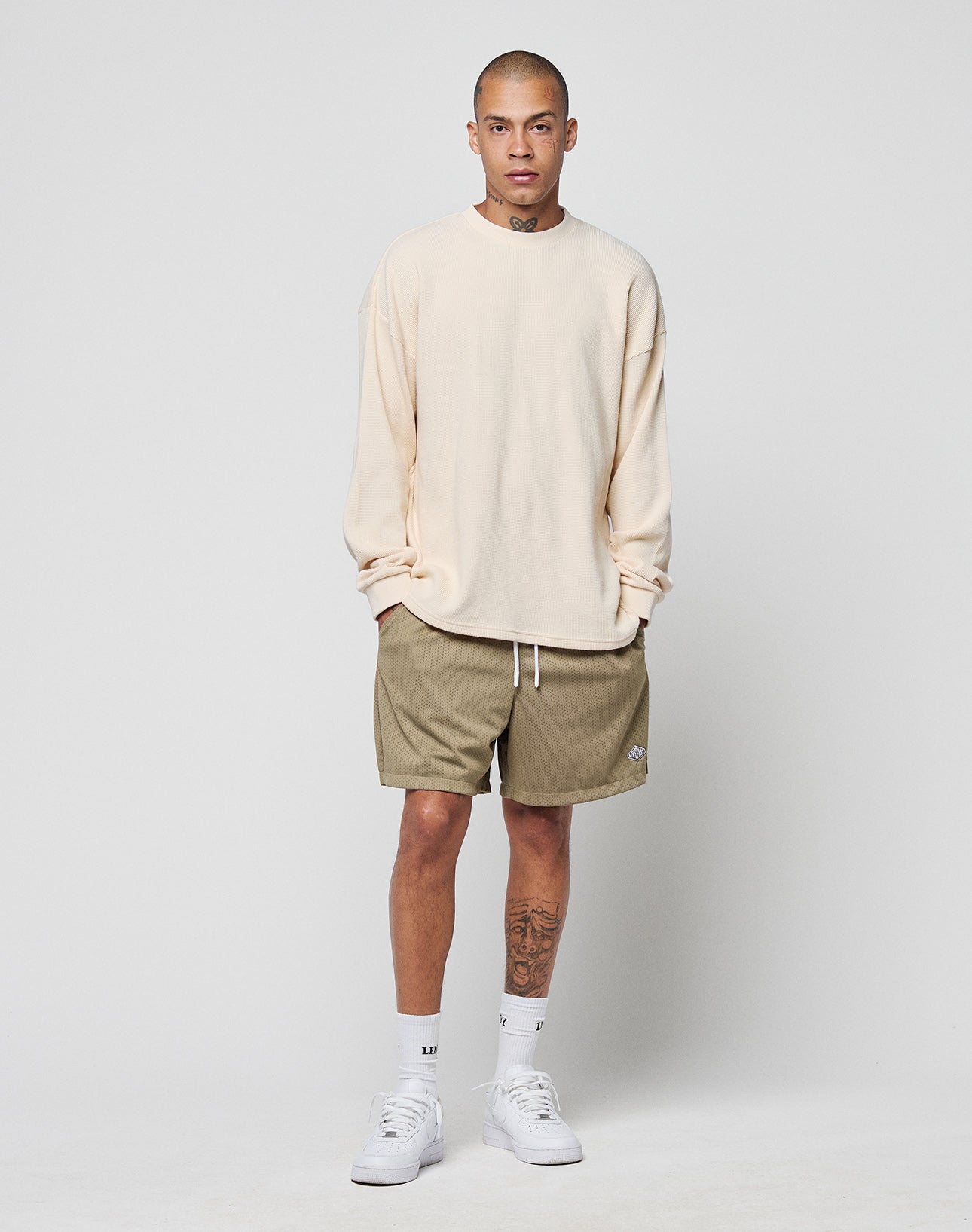 A person with short hair and neck and leg tattoos stands against a plain background, wearing the LIVE FAST DIE YOUNG Blank Waffle Longsleeve—a cream, oversize 100% Baumwolle shirt—olive shorts, white socks, and white sneakers.