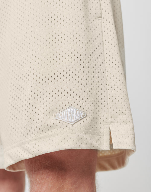 Close-up of a person wearing LIVE FAST DIE YOUNG Basic Mesh Shorts in light beige polyester. The shorts have a regular fit and feature a small white hem logo patch. The persons leg is partly visible below the shorts.