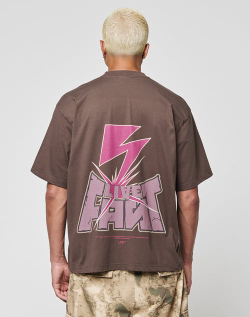 A person with short bleached blond hair is seen from behind, wearing the LFDY Flash Tee by LIVE FAST DIE YOUNG—an oversized brown cotton-slab shirt with a bold pink lightning bolt and LIVE FAST backprint—paired with tan camo shorts.