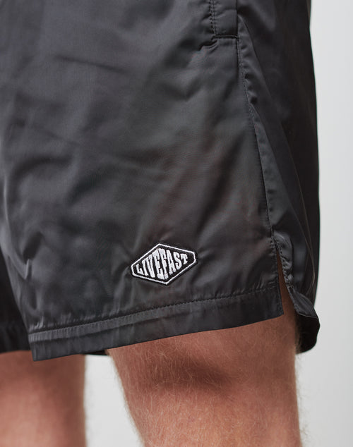 Close-up of a person wearing black Basic Satin Shorts by LIVE FAST DIE YOUNG, featuring an elastic waistband and a small logo patch near the hem. Only the lower part of the shorts and upper thigh are visible.
