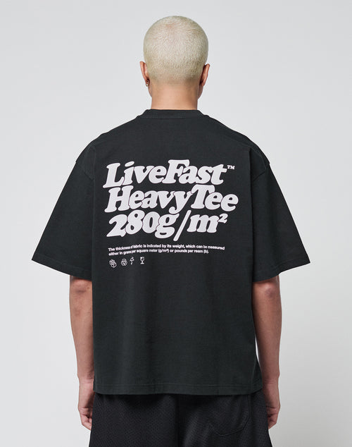 A person with short bleached hair wears a black LIVE FAST DIE YOUNG Basic 280 Tee aus Baumwolle, showcasing bold white “Live Fast Heavy Tee 280g/m²” text with Front- und Backprint icons. The background is plain and light-colored.