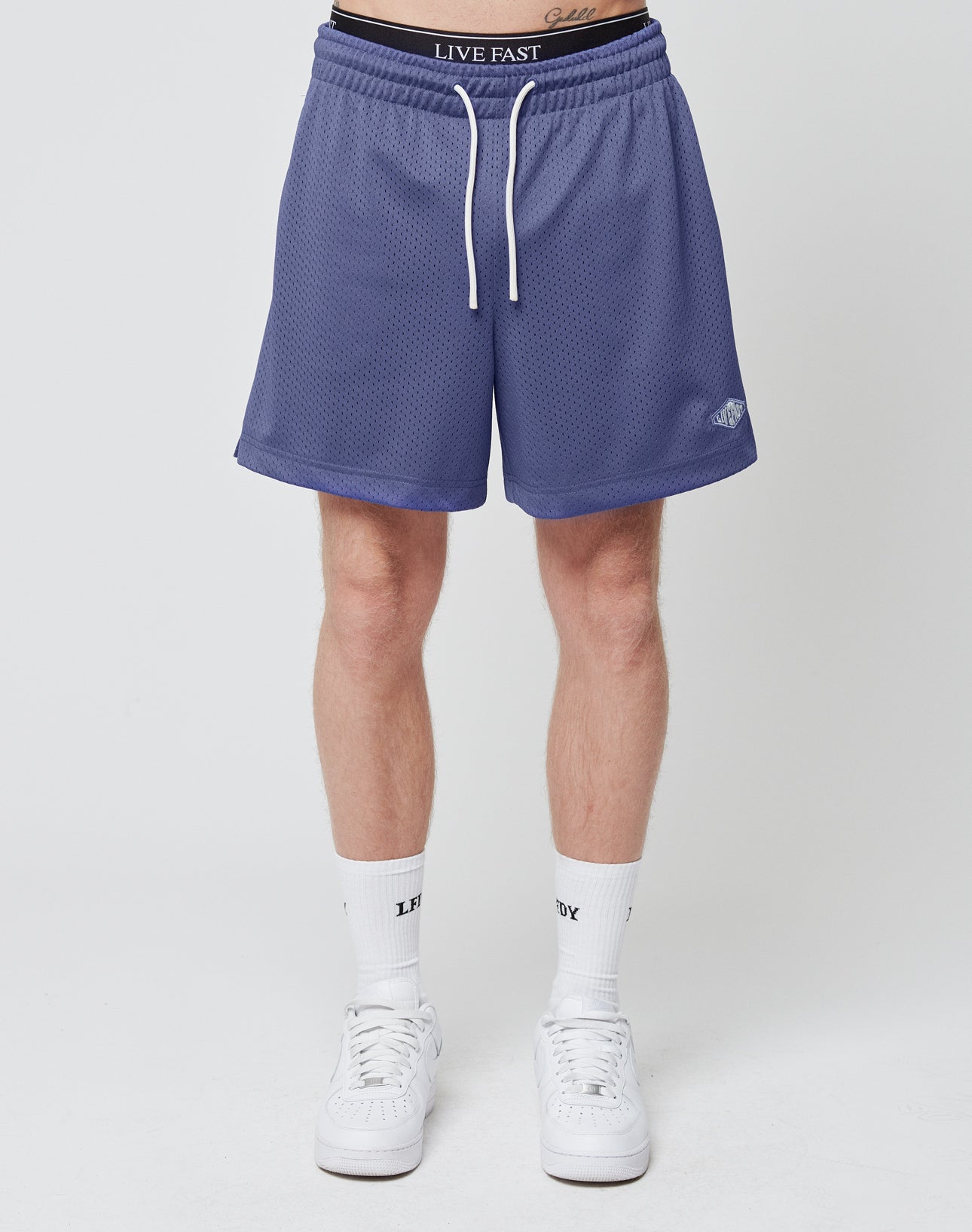 A person wears LIVE FAST DIE YOUNG Basic Mesh Shorts with an elastic waistband and subtle logo patch, paired with white high socks and sneakers, standing against a plain white background. Only their lower torso and legs are shown.