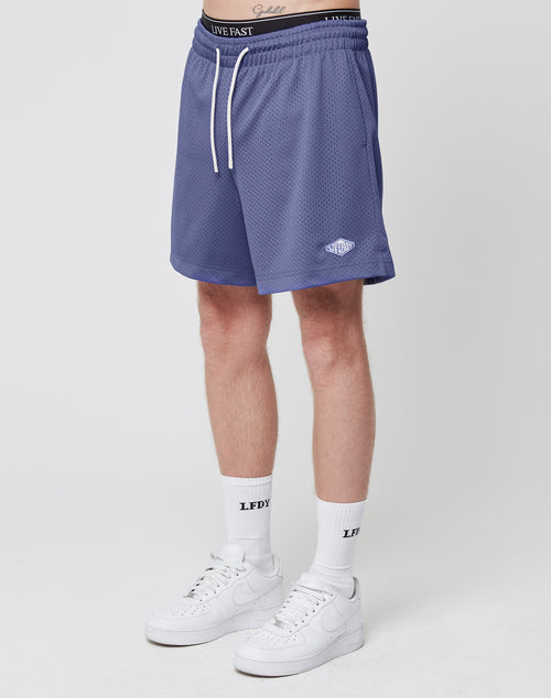 A person wearing LIVE FAST DIE YOUNG Basic Mesh Shorts in blue with an elastic waistband and regular fit, white LFDY socks, and white Nike sneakers stands against a plain white background. Only the lower body is shown.