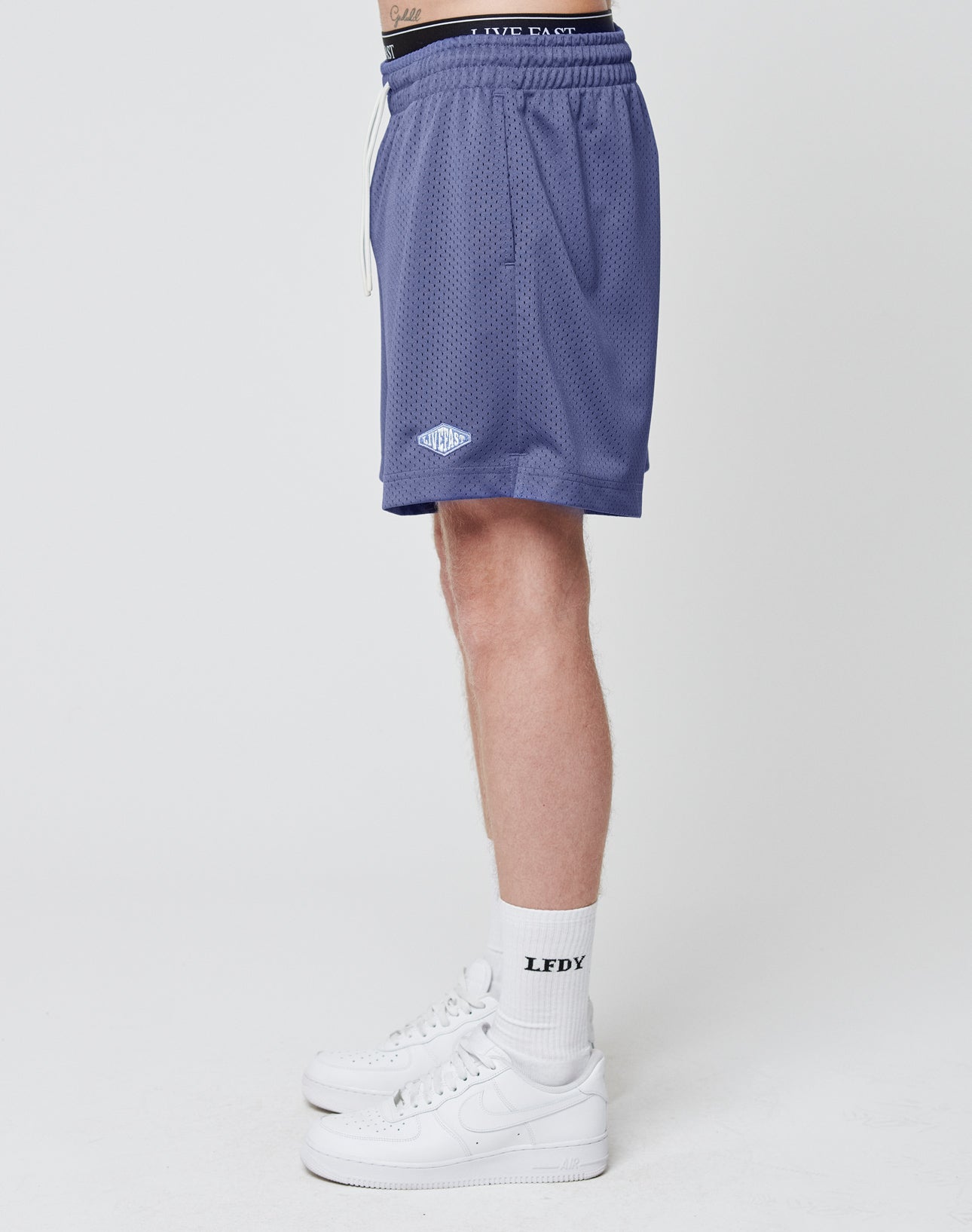 A person is seen from the waist down wearing LIVE FAST DIE YOUNG Basic Mesh Shorts in blue with elastic waistband and regular fit, LFDY-branded white crew socks, and white sneakers, standing sideways against a plain white background.