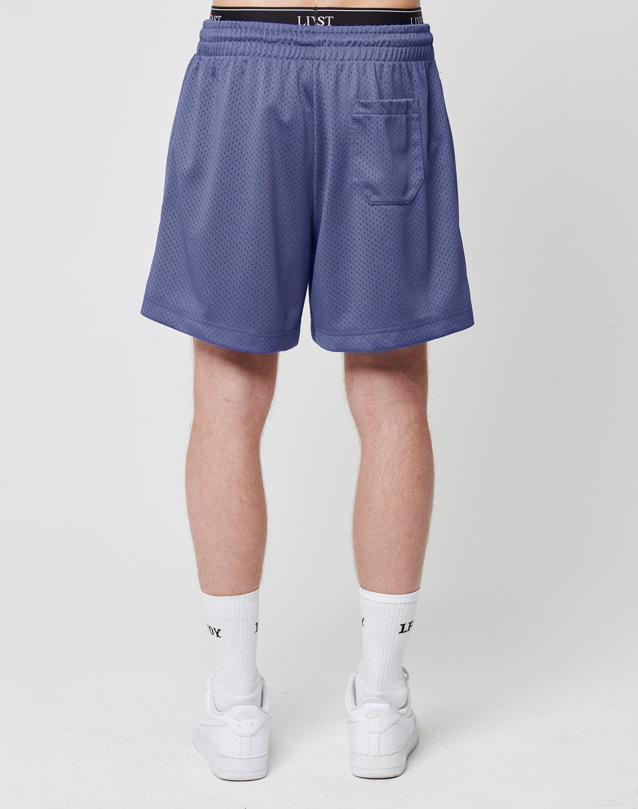 A person faces away, wearing LIVE FAST DIE YOUNG Basic Mesh Shorts in lavender with an elastic waistband and logo patch, plus a small back pocket. They pair them with white crew socks and sneakers against a plain white background.