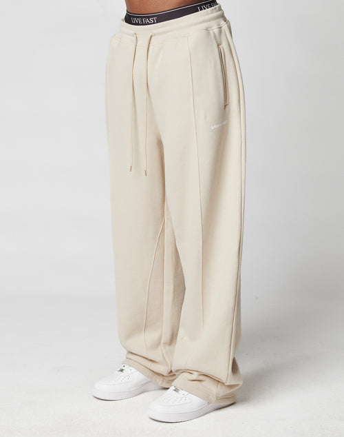 A person wears LFDY Pleated Jogger sweatpants by LIVE FAST DIE YOUNG—loose-fitting, high-waisted, beige with wide fit, elastic waistband, drawstring, side pockets, and “LIVE EAST” on the waist—paired with white sneakers on a white background.