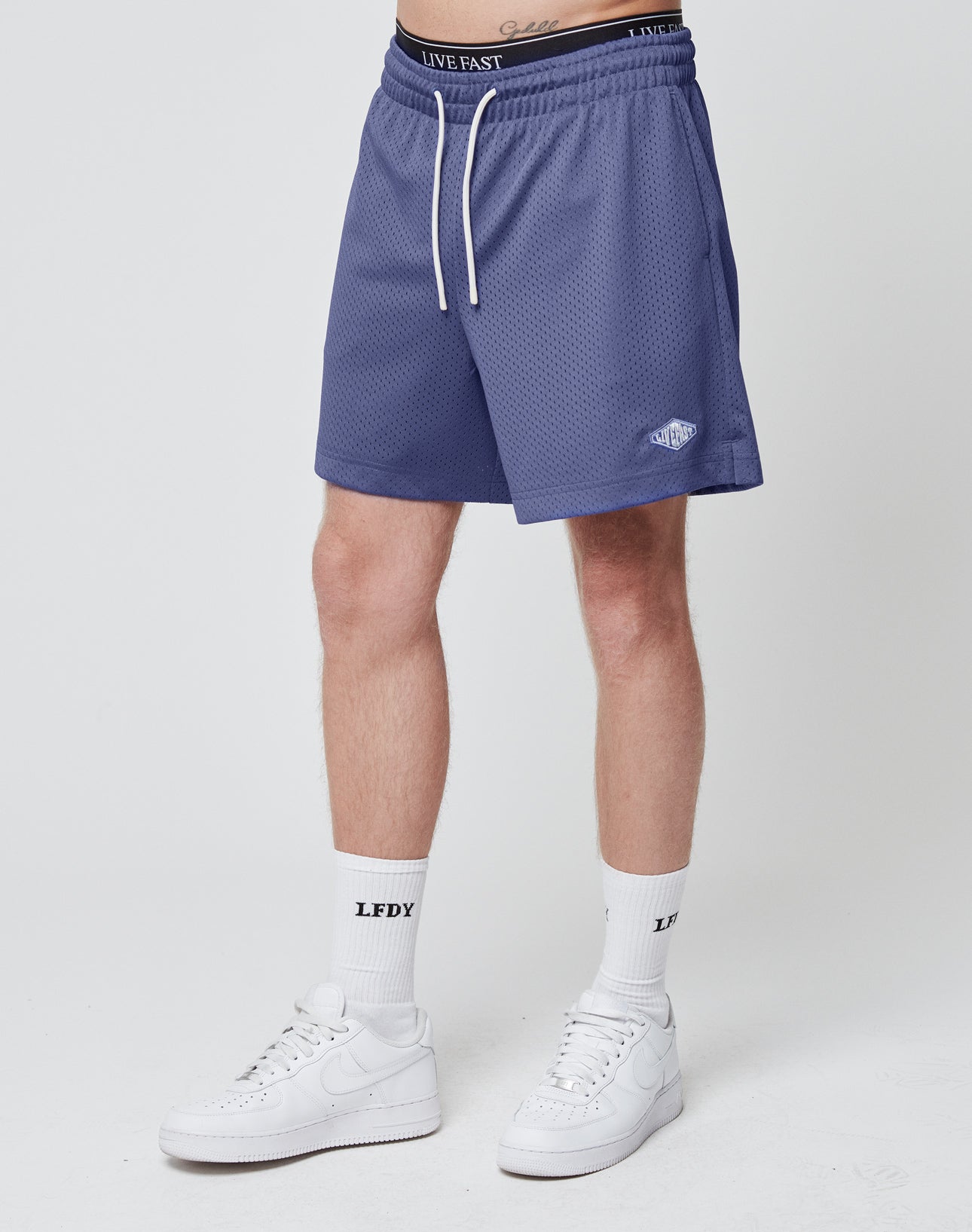 A person wears LIVE FAST DIE YOUNG Basic Mesh Shorts with an elastic waistband and logo patch in purple, paired with white LEDY socks and Nike sneakers. Only their lower body is shown against a plain white background.