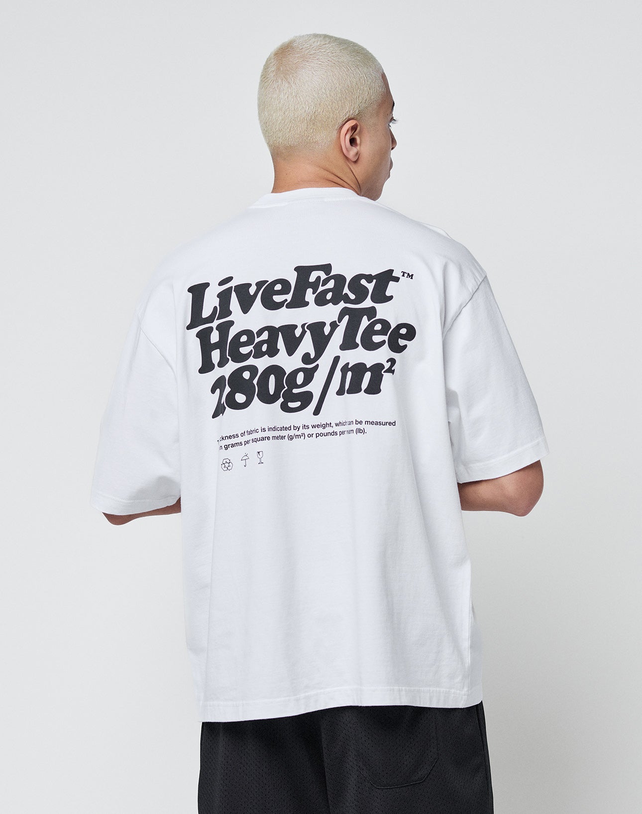 A person with short blond hair wears a white oversize Basic 280 Tee by LIVE FAST DIE YOUNG, featuring bold black backprint text and smaller text below, made from 100% cotton, standing against a plain white background.