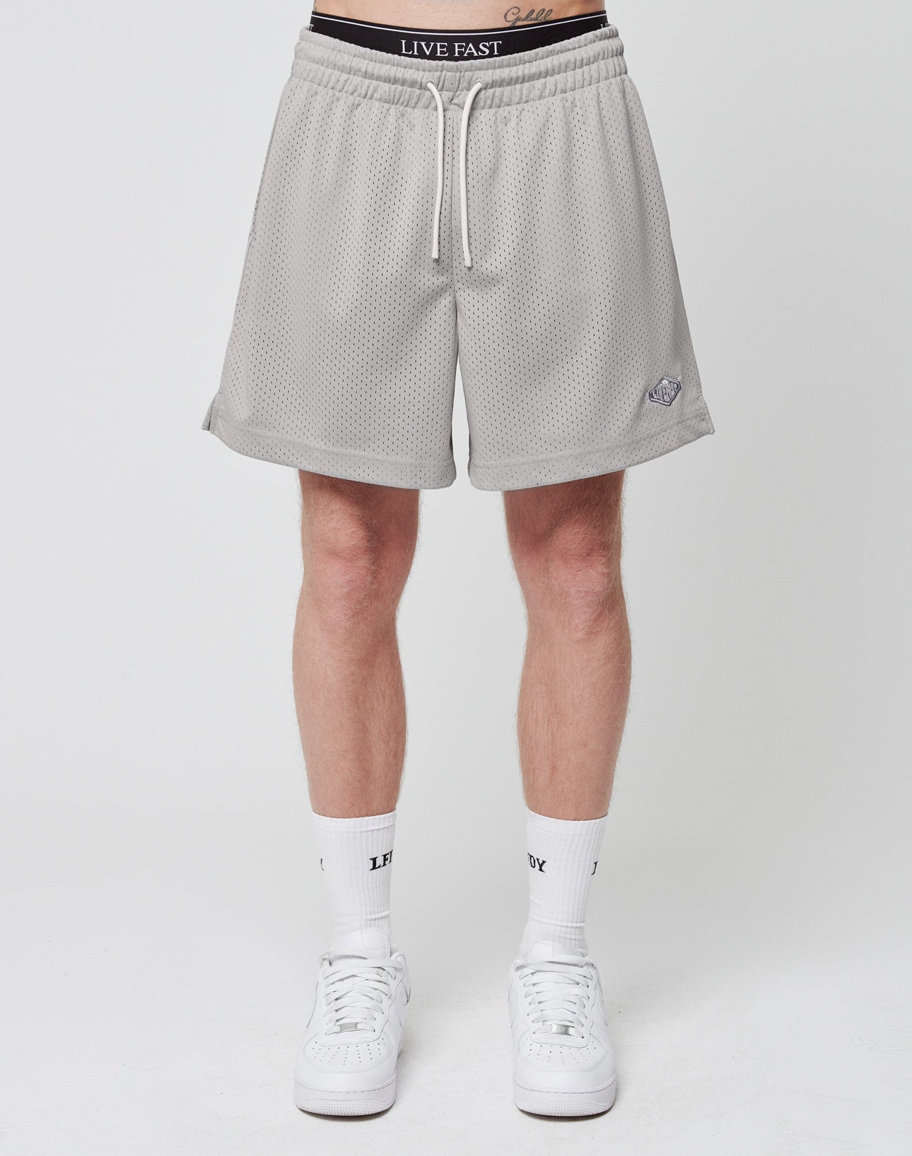 A person stands against a plain background in LIVE FAST DIE YOUNG Basic Mesh Shorts, light gray with a logo patch and white drawstrings at the waist. Made from 100% polyester, theyre styled with white socks and sneakers; LIVE FAST is visible on waistband and socks.