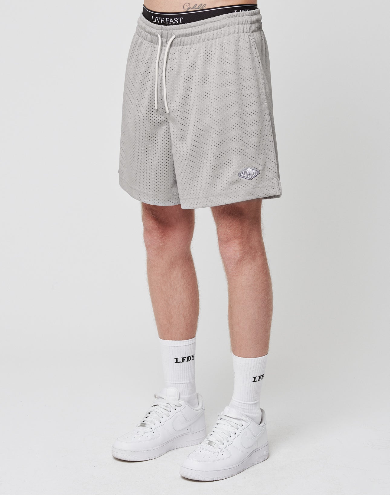 A person wearing LIVE FAST DIE YOUNG Basic Mesh Shorts in light gray with a logo patch, white LIVE FAST boxer waistband, white LFDY socks, and white Nike sneakers stands against a plain white background.