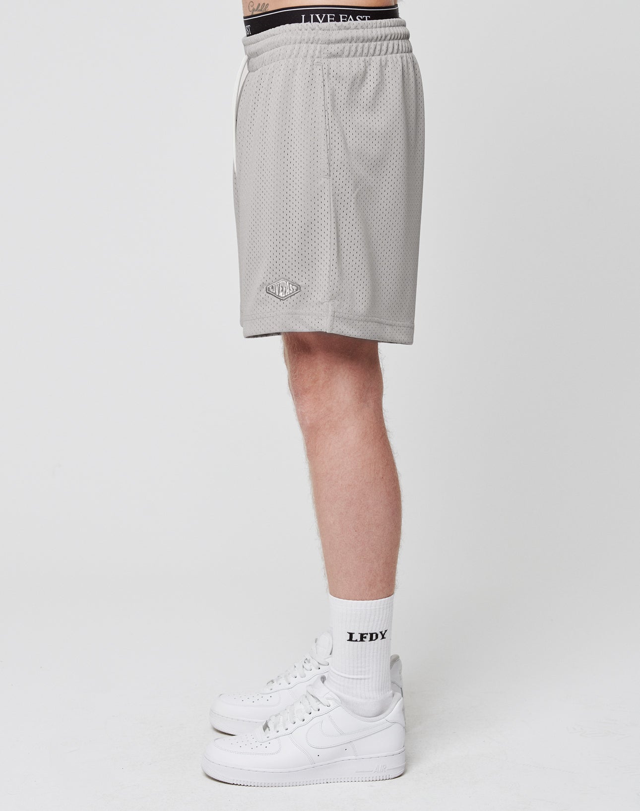 A person stands in profile against a plain white background wearing LIVE FAST DIE YOUNG Basic Mesh Shorts, white crew socks with LFDX text, and white Nike sneakers.