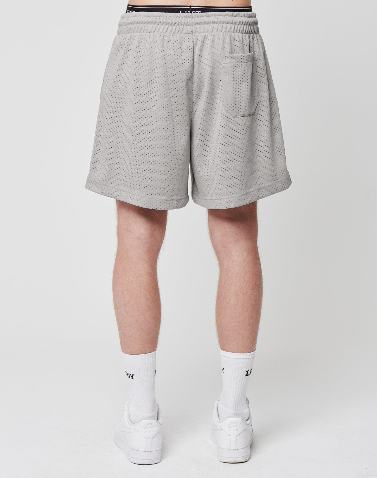 A person faces away, wearing LIVE FAST DIE YOUNG Basic Mesh Shorts in light gray (100% polyester, regular fit), featuring a logo patch, elastic waistband, and small back pocket. Paired with white crew socks and sneakers. Plain white background.